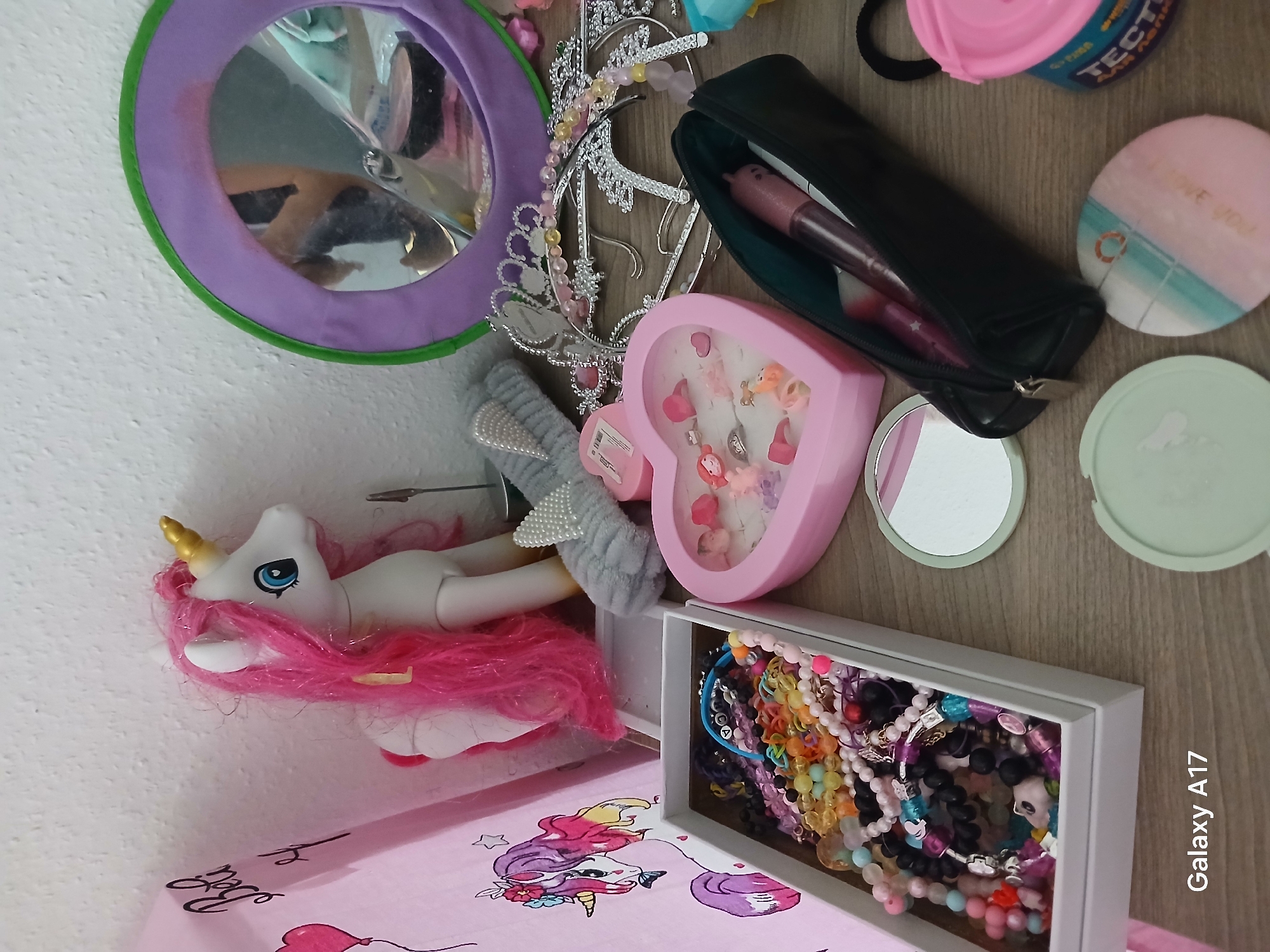 This image shows a collection of children's items on a pink surface with a unicorn illustration. There is a toy unicorn with pink hair, a mirror with a purple and green frame, a tiara, a black pencil case with a pink marker inside, a pink heart-shaped jewelry box, a small round mirror, and a box filled with colorful bracelets. The photograph has "Galaxy A17" watermarked at the bottom right. The location in Yekaterinburg, Russia, is not discernible from the image.