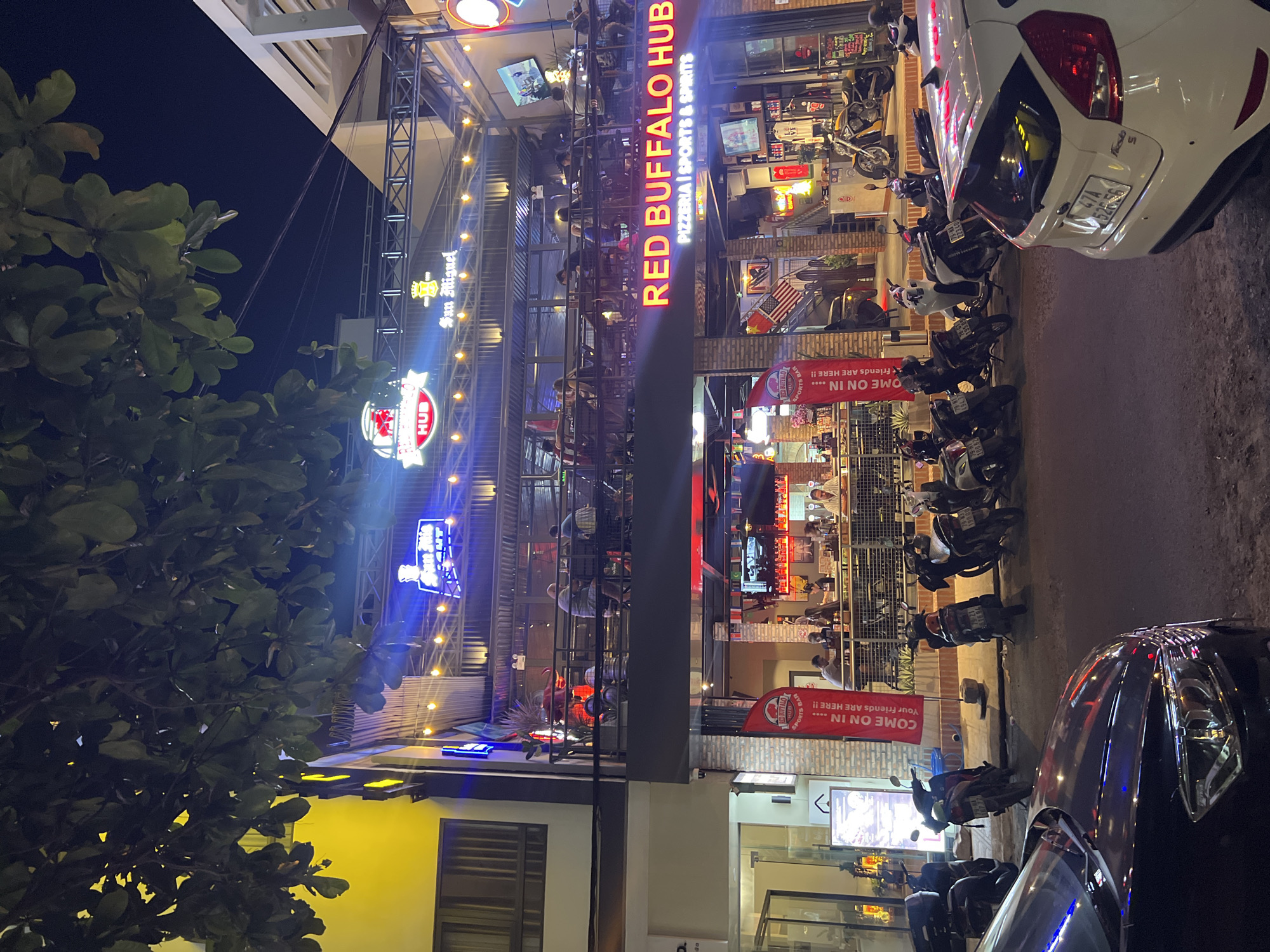 At night in Bắc Nha Trang, Vietnam, a multi-level open-air restaurant called "Red Buffalo Hub Pizzeria | Sports & Spirits" is illuminated with strings of lights and neon signs. Customers are seated on various levels, visible through the open structure. Parked motorcycles line the sidewalk outside the establishment. A white car is parked at an angle to the curb, partially blocking the view, while a dark car occupies the lower right corner of the frame. A tree with dense foliage is on the left side. The building next door has a yellow facade with a window visible. Various signs and banners promote the restaurant, including "Come On In... Your Friends Are Here!!"