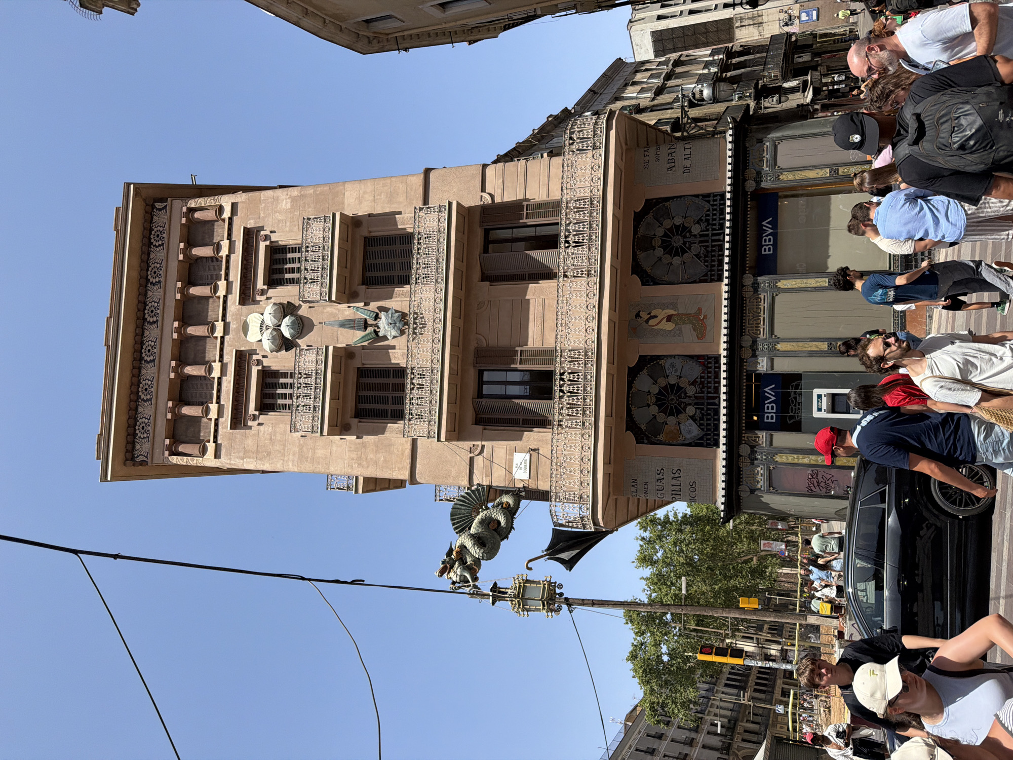The image captures a street scene in Barcelona, Spain, focusing on the ornate facade of a building, likely a commercial establishment, with a distinctive dragon sculpture attached to a lamppost on its corner. The building itself is multi-story, with a light brown facade adorned with decorative elements, balconies with intricate ironwork, and several windows. On the lower part of the building, there are large decorative circular mosaics and what appears to be a storefront with the branding "BBVA".
A diverse group of people are visible on the sidewalk and street. Some are walking, others are standing and conversing, and a few are looking towards the camera or the building. A black SUV is parked on the street in the foreground, and traffic lights are visible in the background. The sky is clear and blue, suggesting a sunny day. The architecture and the presence of the dragon sculpture are characteristic of Barcelona's unique artistic and historical style, possibly hinting at a location in the Gothic Quarter or Eixample district. The dragon, specifically, is a known architectural feature in Barcelona.
