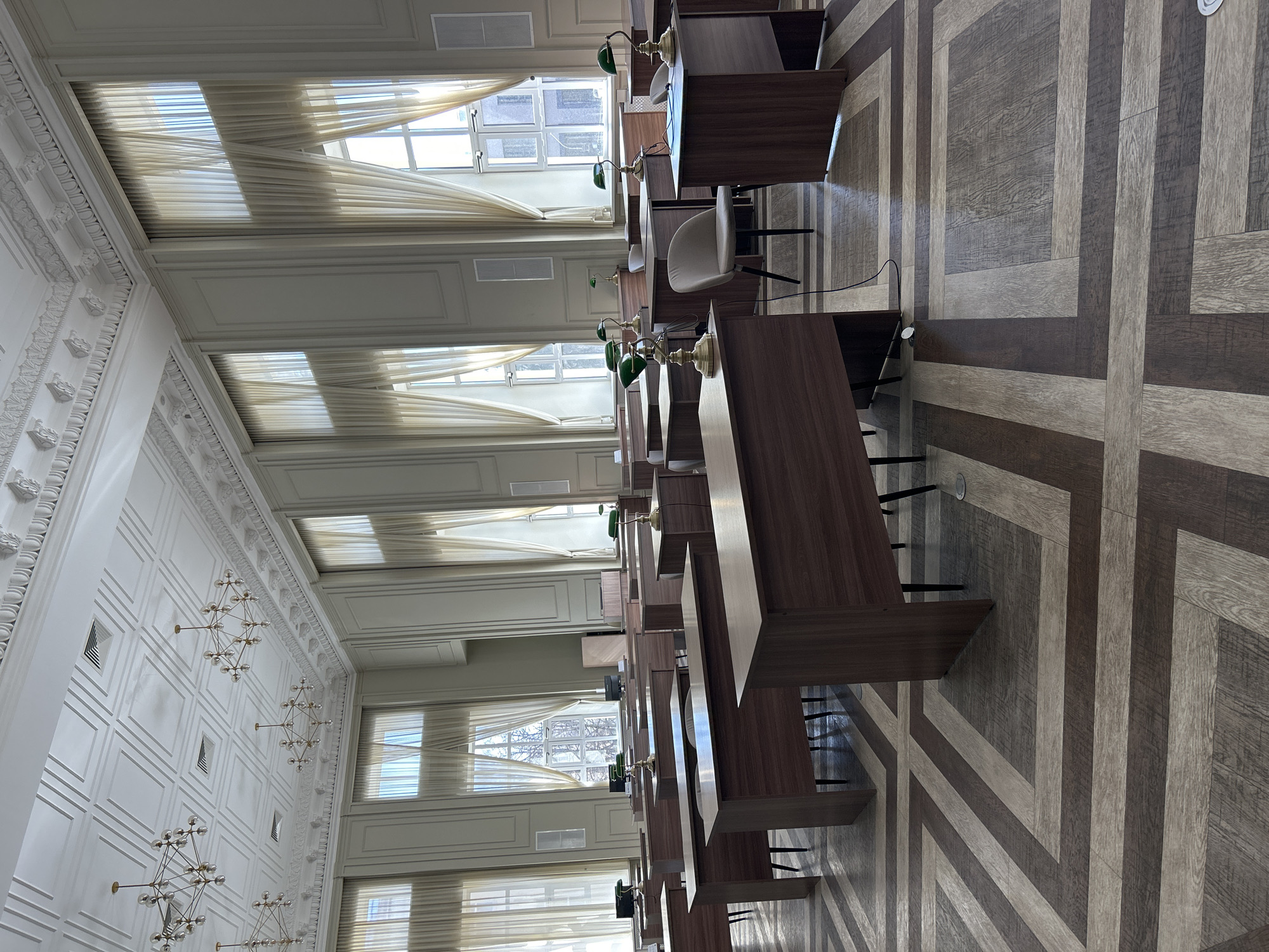 The image shows an interior view of a library or study hall. The location is likely in Izhevsk, Russia, based on the architectural style and typical library setups in that region. There are rows of wooden desks, each equipped with a lamp. Chairs are placed at some desks, indicating usage. Tall windows with curtains are on the left, allowing natural light into the room. The floor has a geometric pattern. No people are visible in the shot. The environment appears calm and organized.