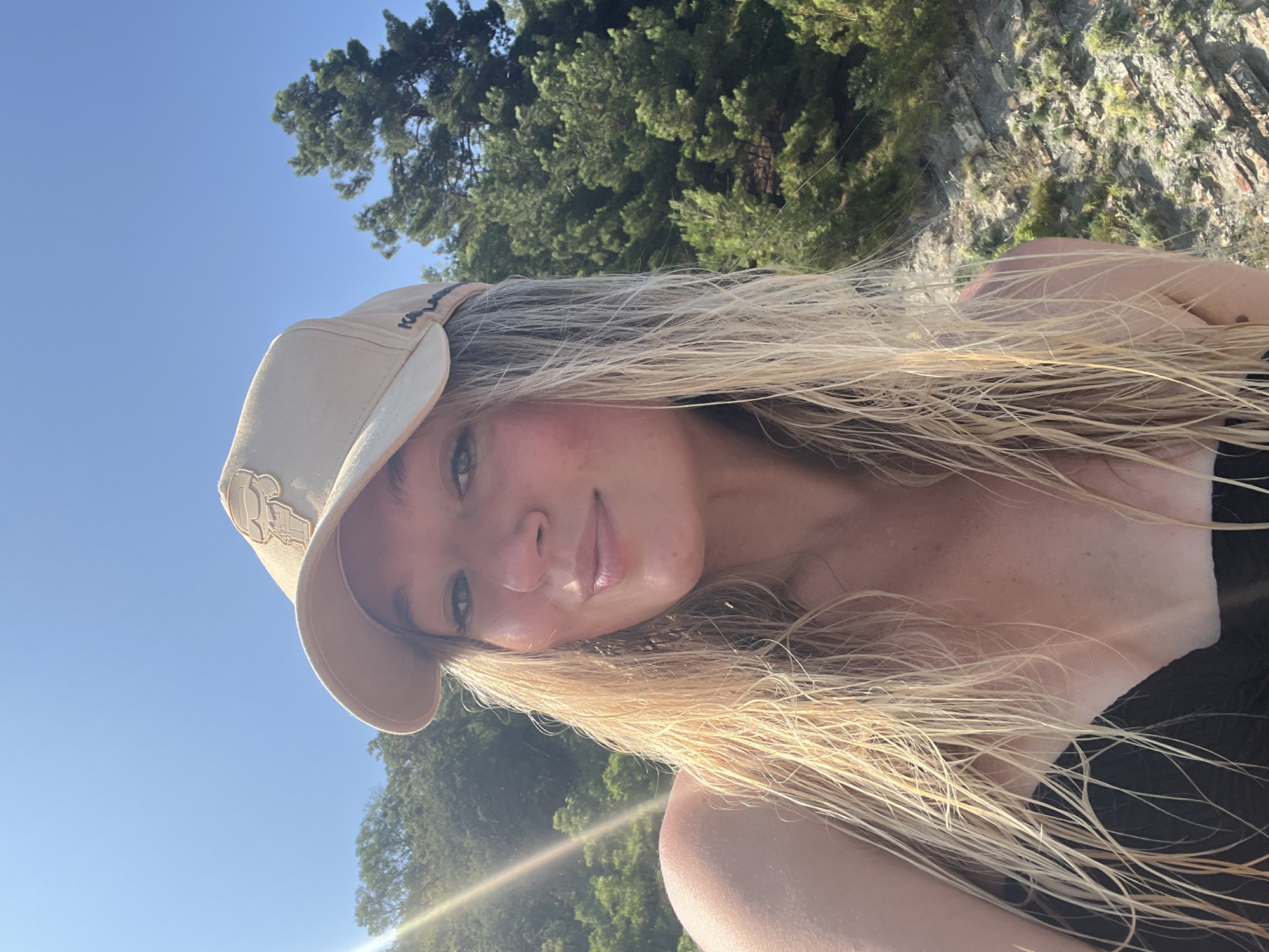 A young woman with blonde, wet hair and a light-colored baseball cap is taking a selfie outdoors. She is wearing a black top. The background consists of green trees and a rocky cliff face. The sun is shining brightly, creating a lens flare on the left side of the image. The overall setting suggests a natural, outdoor location, possibly near the sea or a body of water, which is consistent with the geography of Tuapse, Russia, a city known for its coastal location.