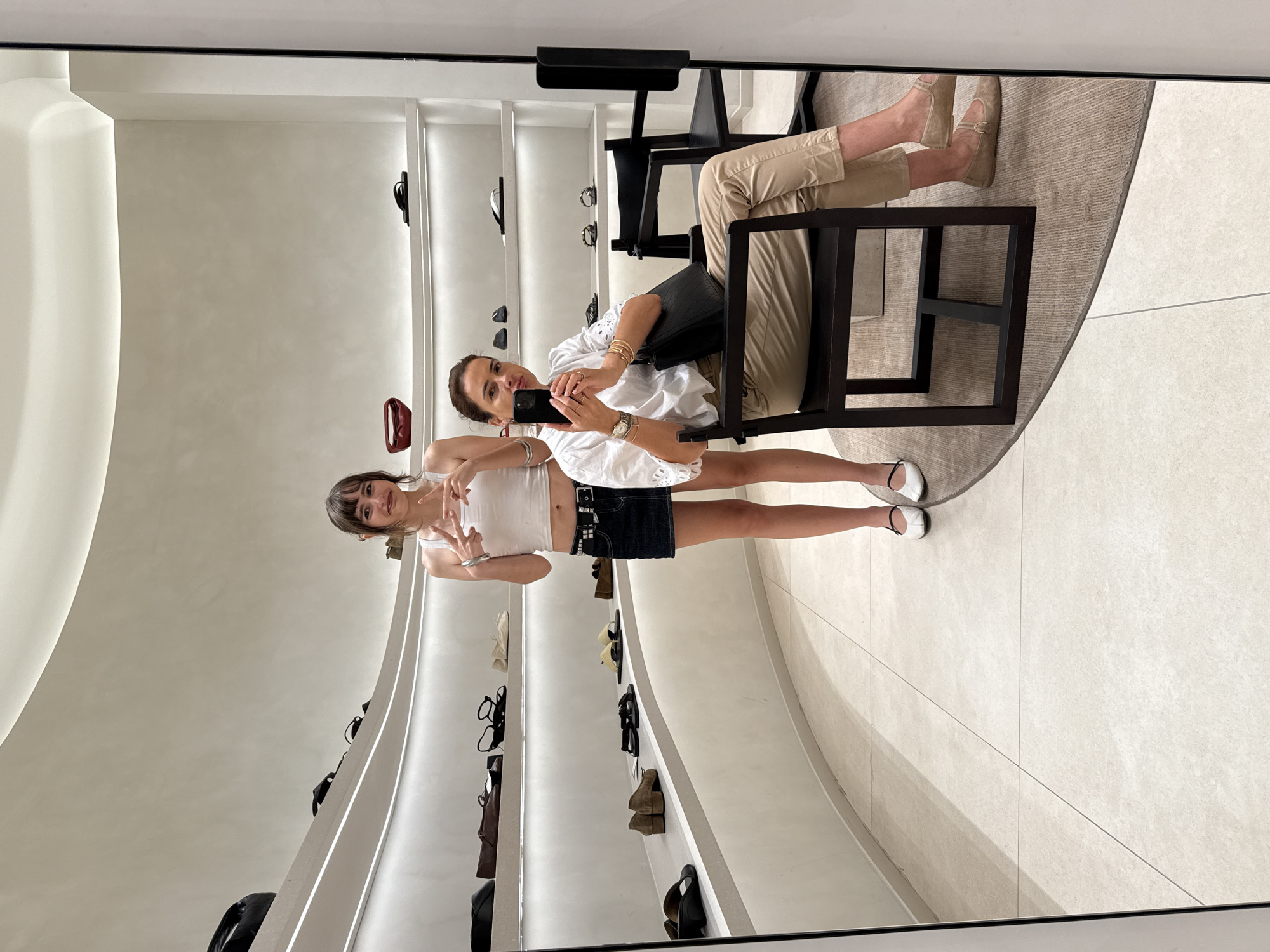 Two women are reflected in a large mirror in a retail setting. The woman closest to the mirror is holding a smartphone and appearing to take a photograph of herself and the other woman. She is wearing a white eyelet blouse, tan slacks, and slip-on shoes and is seated in a dark-colored chair. The younger woman is wearing a white tank top and a dark denim skirt, and is standing giving a peace sign. The retail space has curved white walls with recessed shelving displaying shoes and handbags. The floor is tiled in a light color and a circular rug sits beneath the chair. A portion of the ceiling is visible at the top of the image. The image does not provide enough information to confirm that this is in Loures, Portugal.