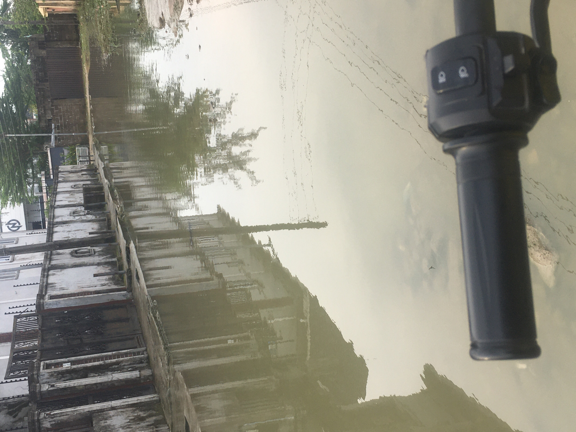 A low-angle, forward-facing, full shot captures a flooded street in Iranla, Nigeria. The foreground is dominated by the handle of a motorcycle, complete with its controls. The street itself is submerged in murky, reflective water, mirroring the surrounding buildings and vegetation. On the left, a white wall with black gates and ornate metalwork extends along the flooded sidewalk. Further down the street, a dark fence encloses more properties. The water is so high that it reaches the base of the buildings, suggesting recent or ongoing heavy rainfall.