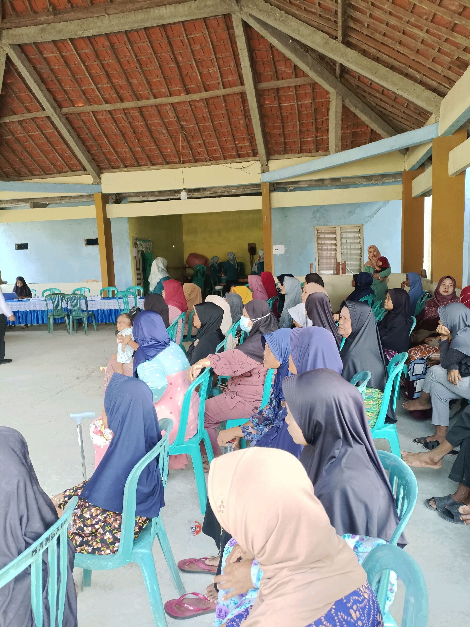 In Rancang Kencono, Indonesia, a group of people are gathered inside a covered outdoor structure with a tiled roof. The structure appears to be a community hall or waiting area. In the foreground, several individuals, predominantly women wearing headscarves, are seated on teal plastic chairs. Some are looking forward, while others are engaged in conversation. A baby is being held by one of the women. In the background, more people are seated, and a table is set up with blue tablecloths and what appear to be registration papers or documents. A person in a white shirt and dark pants stands near the table. The environment is simple, with concrete floors and light-colored walls. The scene suggests a community event or a gathering for a service.