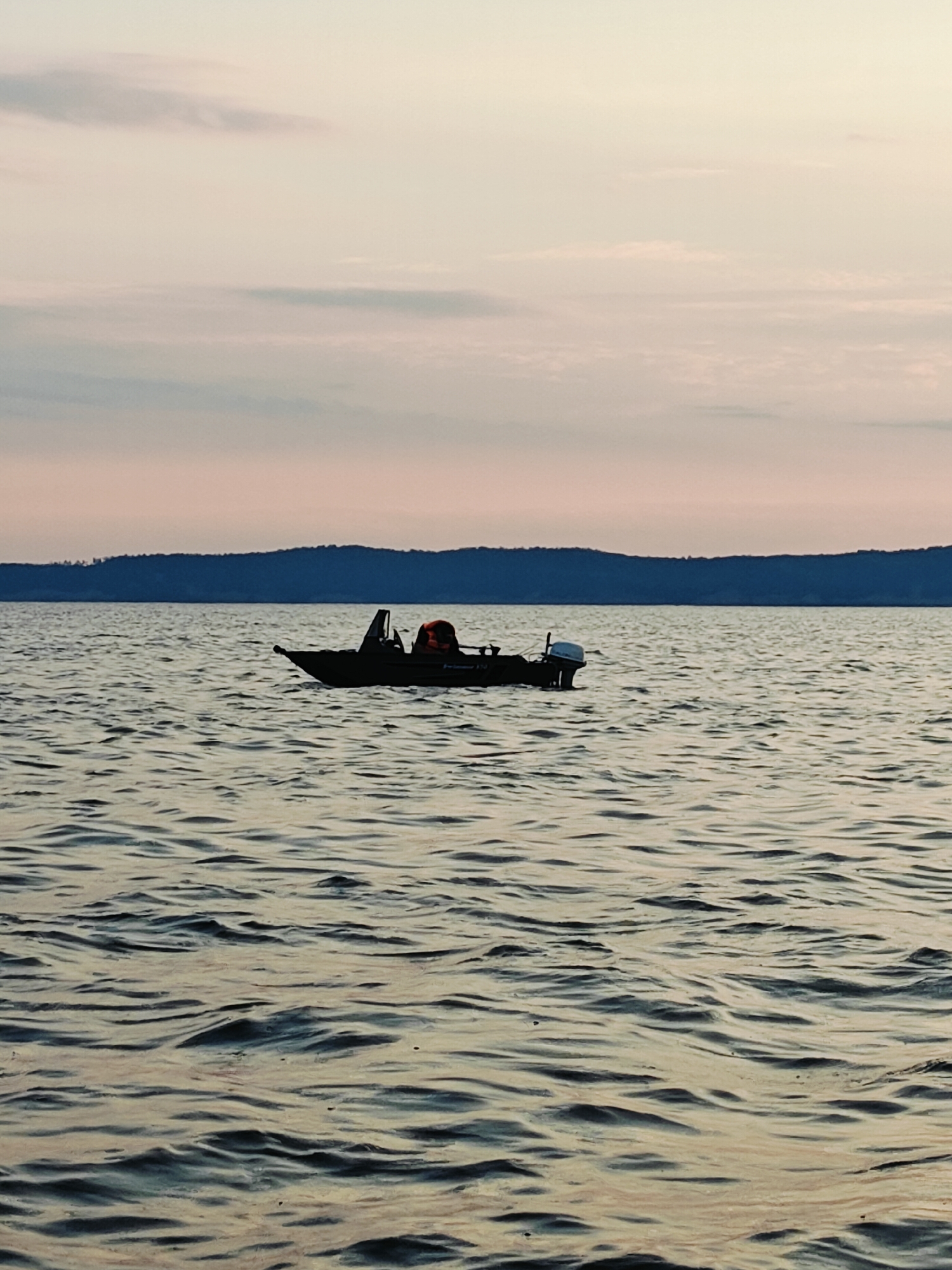 A small, dark motorboat is on the water, seen from a low angle. The boat has an outboard motor on the stern and a person wearing an orange life jacket in the center. The water is choppy, with ripples and waves reflecting the soft colors of the sky, which is a gradient of pale pink, yellow, and blue, with faint clouds. In the background, a dark, wooded shoreline is visible under the horizon line. The scene is likely a body of water near the city of Chaykovsky, Russia, possibly during dawn or dusk due to the lighting.