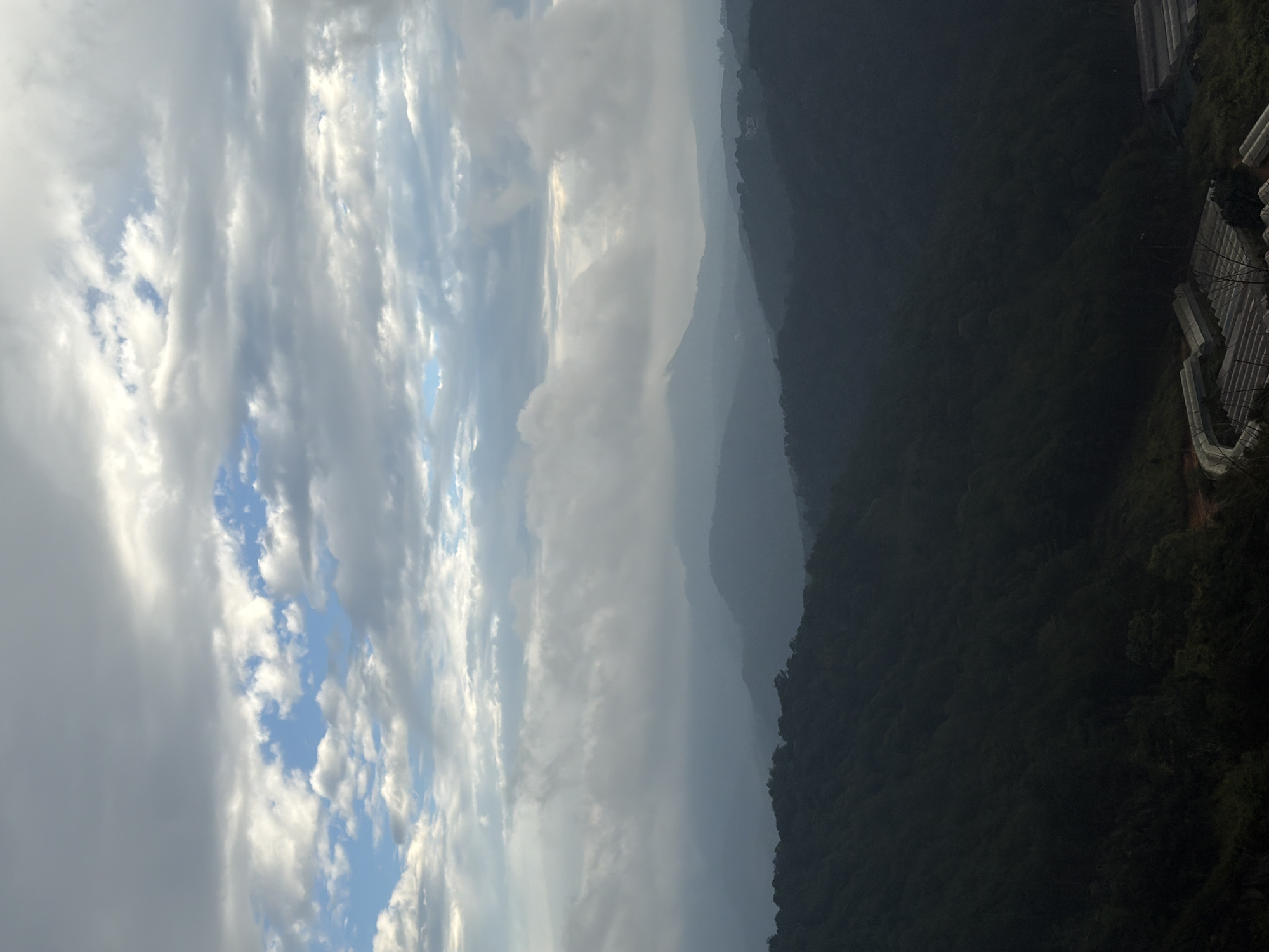 The image shows a panoramic view of rolling hills and mountains, shrouded in mist and clouds. The sky is a mix of gray clouds and patches of blue. The foreground is dominated by dark green, densely forested hills, suggesting a natural, possibly tropical landscape. In the lower right corner, there are structures that appear to be greenhouses or agricultural facilities, indicative of human activity and farming. The overall atmosphere is serene and slightly melancholic, possibly captured at dawn or dusk due to the diffused light. The location, Xuân Thành, Vietnam, is known for its mountainous terrain and agricultural activities, which aligns with the visual elements in the photograph. There are no people visible in the image.