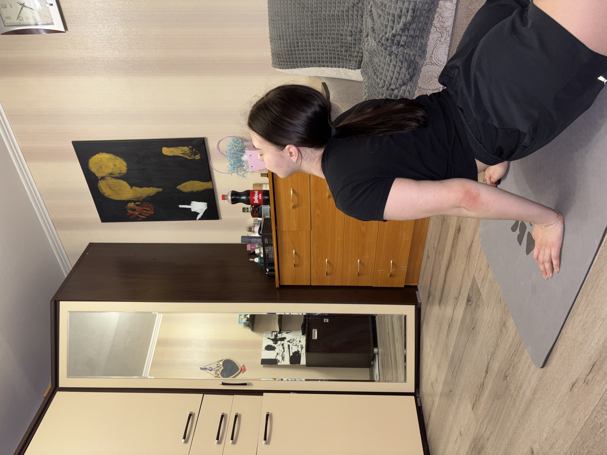 A woman with dark hair in a ponytail is doing a yoga pose on a gray mat. She is wearing a black t-shirt and black shorts. The mat is on a light-colored wood floor. Behind her is a couch with gray cushions and a patterned blanket. A wooden dresser stands against a beige wall, and various objects are on top of it, including a Coca-Cola bottle. On the wall above the dresser is a black-painted canvas, with golden or yellow-colored figures. A wardrobe with a mirror is on the left side of the image. The setting appears to be indoors, in a room in Perm, Russia.