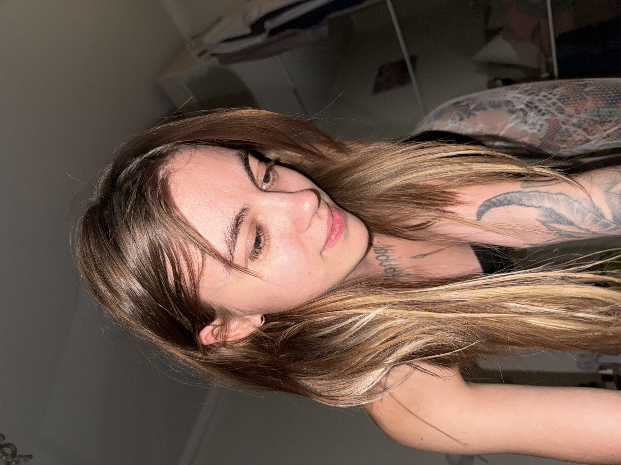 A young woman with long, light brown hair and tattoos on her neck and arms is shown in a close-up, indoor shot. She is looking down and to her right, with her head tilted. The background is blurred and appears to be a domestic setting, possibly a bedroom, with clothing hanging on a rack visible on the right side of the frame. The lighting is dim, creating shadows on her face and body. There is no visible indication of the image being taken in Moscow, Russia.