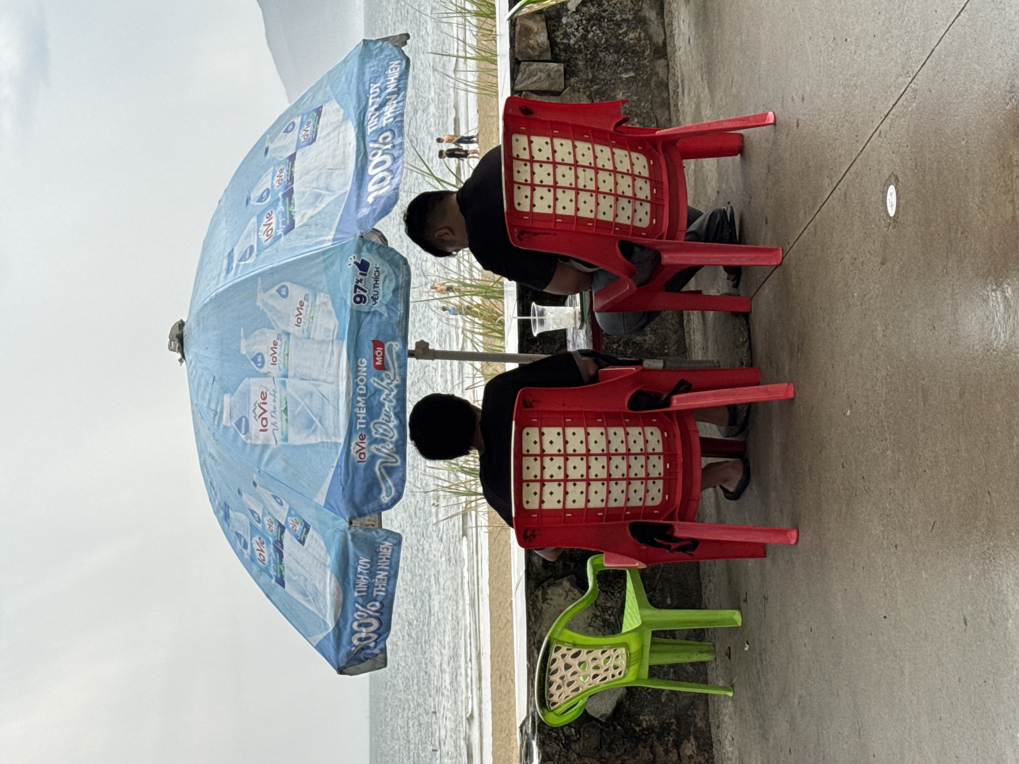 This is an eye-level, medium shot of two men sitting at a table under a blue umbrella on a cloudy day. They are facing away from the camera, with their backs to the viewer. They are sitting on red plastic chairs. The man on the left has a black t-shirt and dark pants, and the man on the right has a black t-shirt and dark jeans. In front of them is a table with a plastic cup and a bottle of water. To the left of the men is a bright green plastic chair. The umbrella is decorated with images of water bottles and text in Vietnamese. In the background, there is a beach with sand and the ocean. Several people are visible on the beach, including some children playing in the water. The image appears to be taken in the city of Đà Nẵng, Vietnam, as indicated by the presence of a La Vie water bottle brand and the beach setting, which is a common sight in coastal Vietnamese cities.