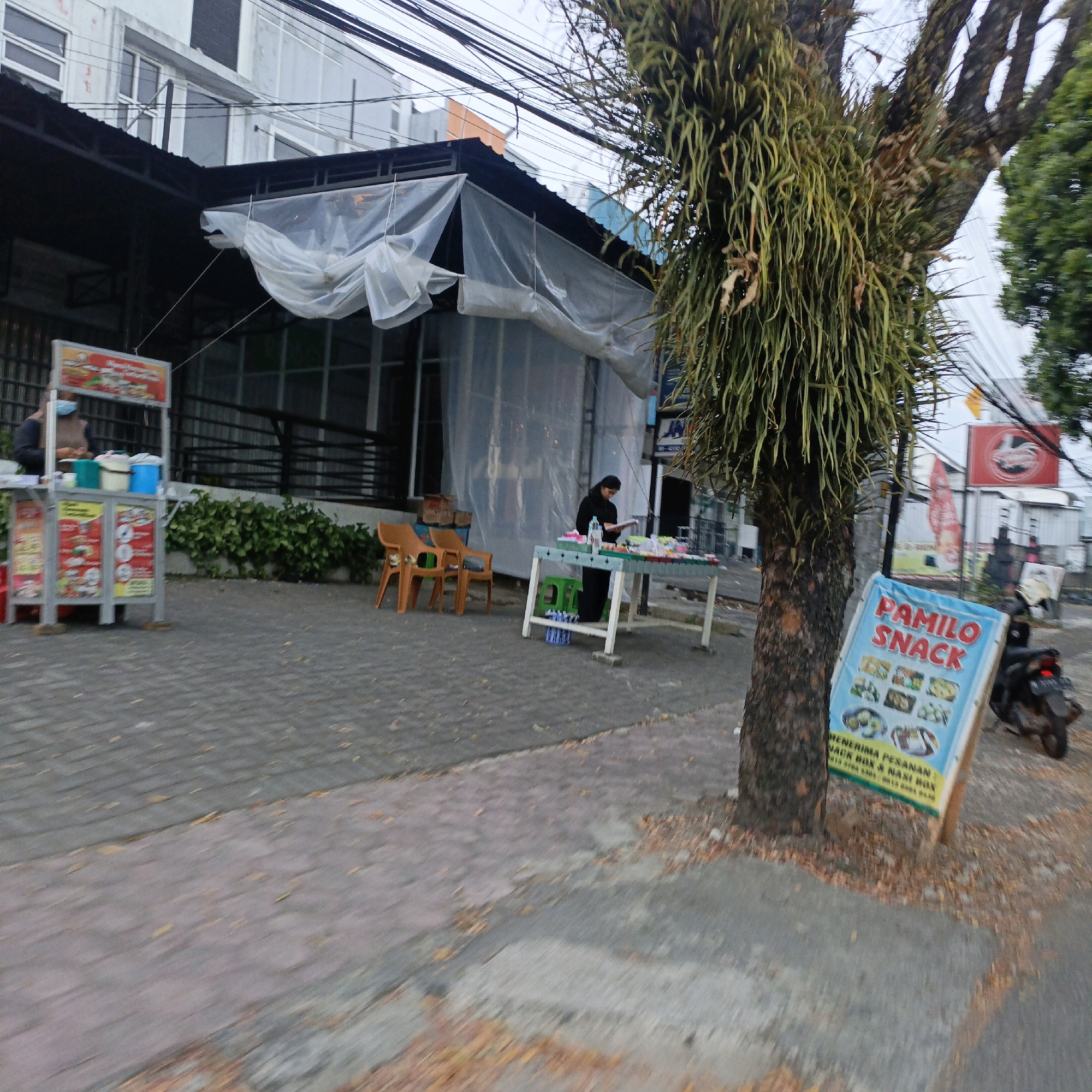 In Salatiga, Indonesia, an outdoor scene depicts a sidewalk next to a building under construction or renovation, indicated by a translucent plastic sheeting covering a portion of the structure. On the sidewalk, a food cart with several colorful display panels featuring images of food is positioned to the left. A person wearing a light-colored shirt and dark pants is visible behind the counter of the cart, interacting with items on display. Further down the sidewalk, a woman in a dark outfit is standing at a table laden with various food items and beverages. She appears to be engaged with the items on the table. Two orange plastic chairs are placed in front of a section of the building, and a green plastic stool is near the table where the woman is standing. A tree with hanging epiphytic plants stands prominently in the foreground to the right, with a sign board in front of it that reads "PAMILO SNACK" and displays images of food items. A parked motorcycle is visible behind the sign. The ground is paved with interlocking bricks and scattered leaves. The overall environment suggests a commercial area with food vendors.