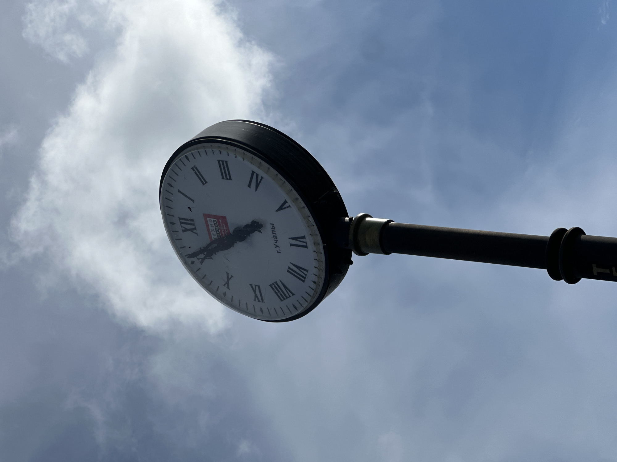 A low-angle shot captures a large, round street clock set against a cloudy, daytime sky. The clock features a white face with black Roman numerals, ranging from I to XII. The clock hands, dark in color, are positioned to indicate approximately 1:30 or 2:30. Some wear or dirt is visible on the clock face, particularly around the center.
On the lower half of the clock face, below the center, the Cyrillic text "г. Учалы" is clearly visible. Near the center, a red rectangular emblem with white text is present, partially obscured by the clock hands. While difficult to read completely, parts of it resemble "ТЕХНО" and "СЕРВИС", possibly indicating a brand or service provider.
The clock is mounted on a thick, dark pole or arm, which extends horizontally into the frame on the right side. The sky in the background displays a mix of light blue patches and soft, scattered white and gray clouds, suggesting a bright but possibly overcast day. The perspective is looking upwards, emphasizing the clock against the vastness of the sky.