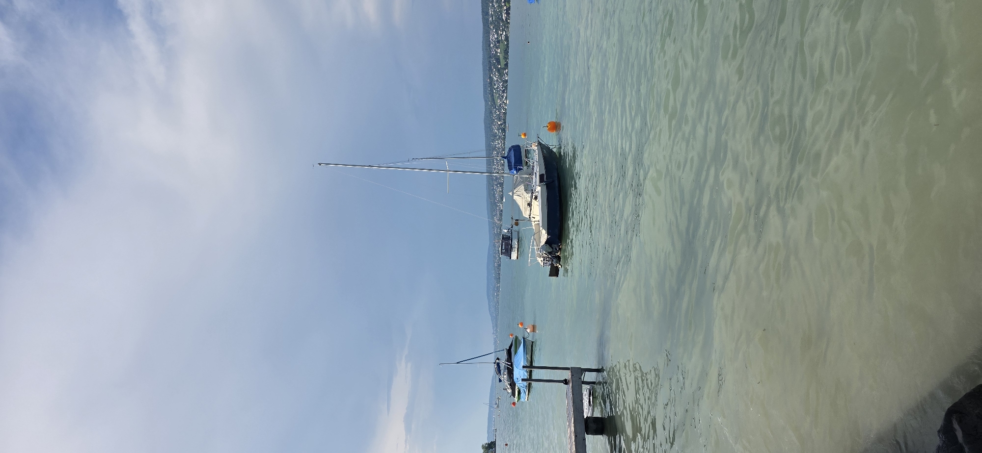 The image depicts a tranquil scene on Lake Zurich, specifically near the municipality of Oberrieden, Switzerland. In the foreground, a wooden pier juts out into the shallow, greenish water. Several boats are visible on the lake. A sailboat with a tall mast is anchored in the mid-ground, while other smaller boats, some with covers, are scattered across the water. In the distance, the shoreline is lined with a picturesque town, its buildings nestled among trees against a backdrop of rolling hills under a cloudy sky. The water is calm, with gentle ripples reflecting the sky. There are no people visible in the image.