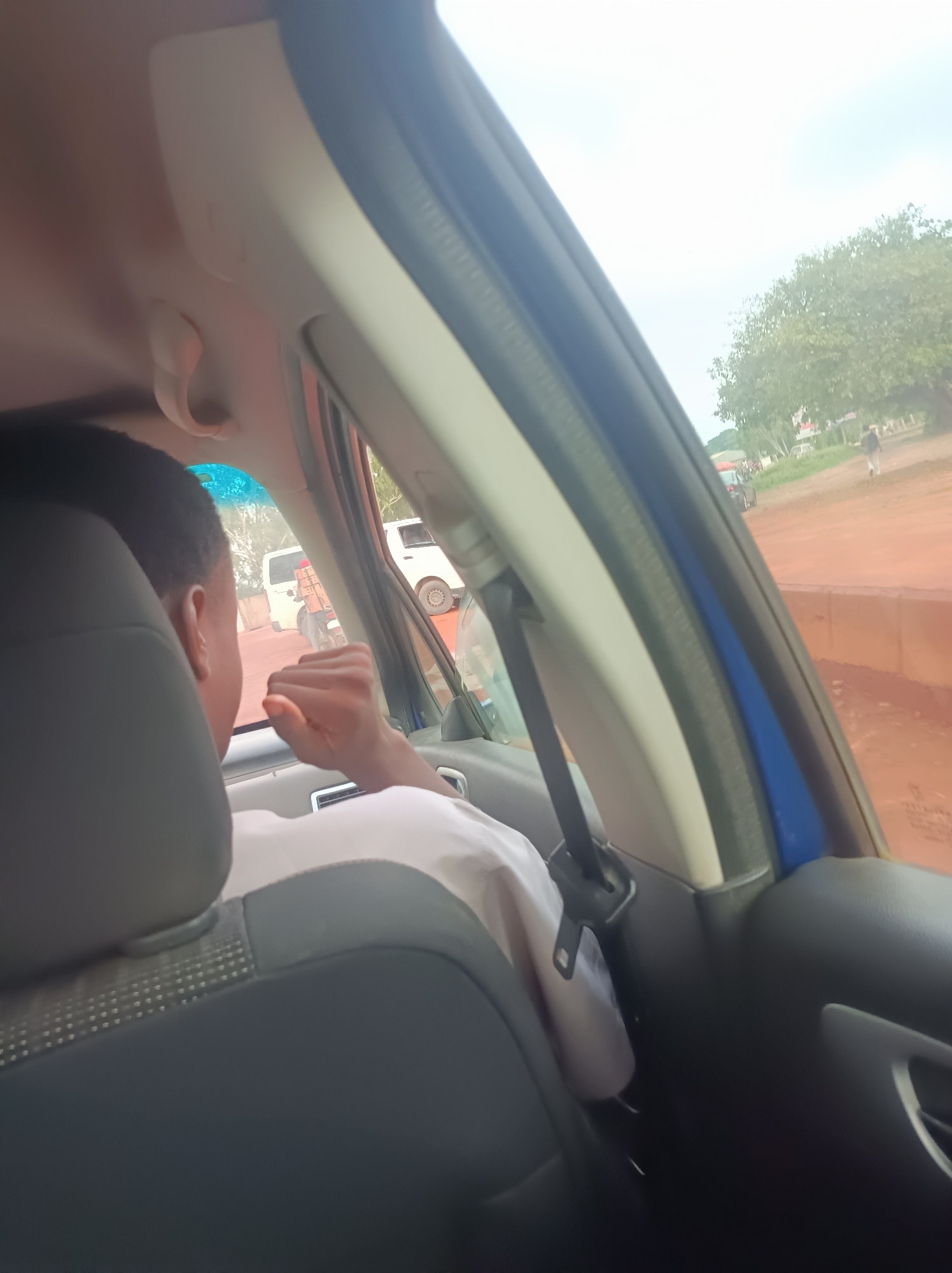 The image presents a view from inside a vehicle, looking out through the front passenger side window. A person with short, dark hair is seated in the front passenger seat, seen from behind, wearing a light-colored, possibly white, short-sleeved shirt. Their right arm is visible, with a lightly clenched hand resting near the window. The car's interior includes a dark grey/black seat with a textured pattern on the backrest, a light-colored overhead panel with a grab handle, and a blue trim along the A-pillar. A black seatbelt is visible, extending down past the person's shoulder.
Outside the car, a reddish-brown dirt road is visible, characteristic of unpaved or local roads, consistent with the Barnawa, Nigeria context. Along the road, various elements appear: a white van, a person on a motorcycle, and another person walking further in the distance. The roadside features green trees and what appear to be buildings beyond them. The sky is bright and overcast, suggesting daytime conditions with diffused light. The scene depicts typical activity along a road, implying the car is either moving or stopped in traffic. No discernible text is visible.