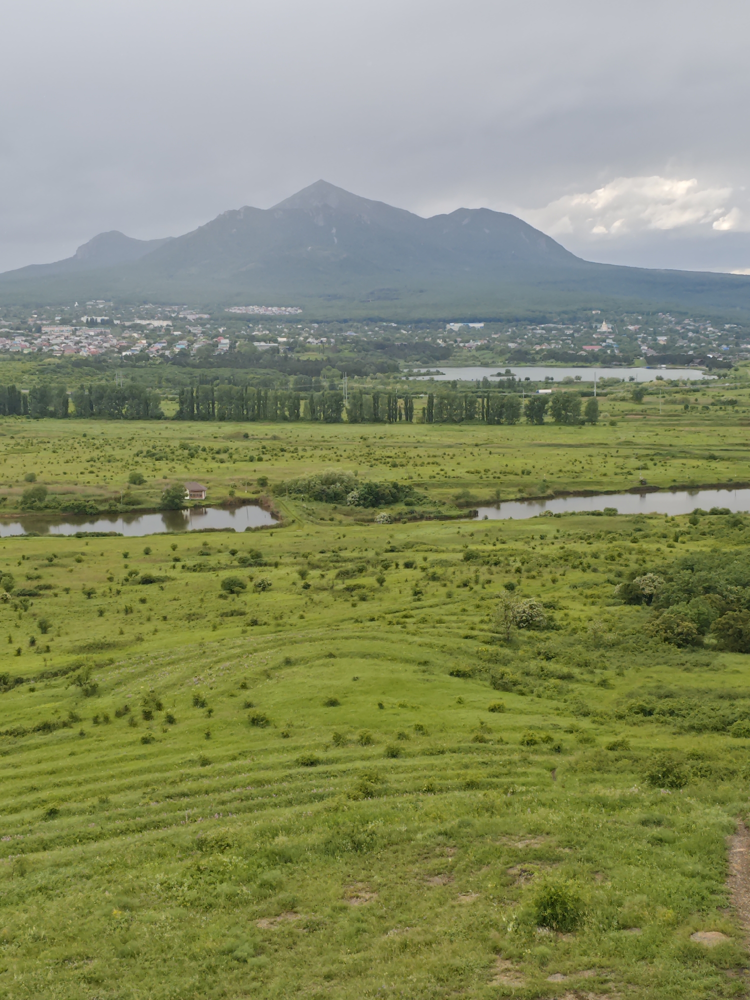 The image shows a wide, grassy landscape in Inozemtsevo, Russia, with a winding river in the mid-ground. In the distance is a town, beyond which rises a large mountain. The sky is overcast.