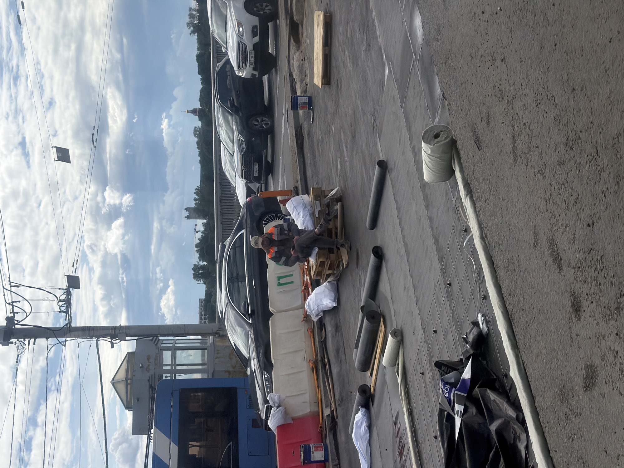In an outdoor urban setting, possibly a construction site or street repair area in Saint Petersburg, Russia, a person wearing work clothes, including an orange safety vest, sits on a wooden pallet. The individual appears to be taking a break from work. The scene features several cars parked nearby, and various construction materials such as white and red plastic barriers, rolled-up materials that appear to be insulation or roofing, and large diameter pipes. A blue vehicle, possibly a bus, is partially visible on the left. The sky is partly cloudy with a mix of white clouds and blue sky, suggesting it could be daytime with variable weather. A roll of material, possibly tape or a sealant, is mounted on a pole or structure in the foreground, with a roll of similar material placed on the ground. Some black plastic sheeting and bags are also present on the ground. The overall impression is a scene of ongoing work in an urban environment. There is no visible text that can be clearly deciphered.