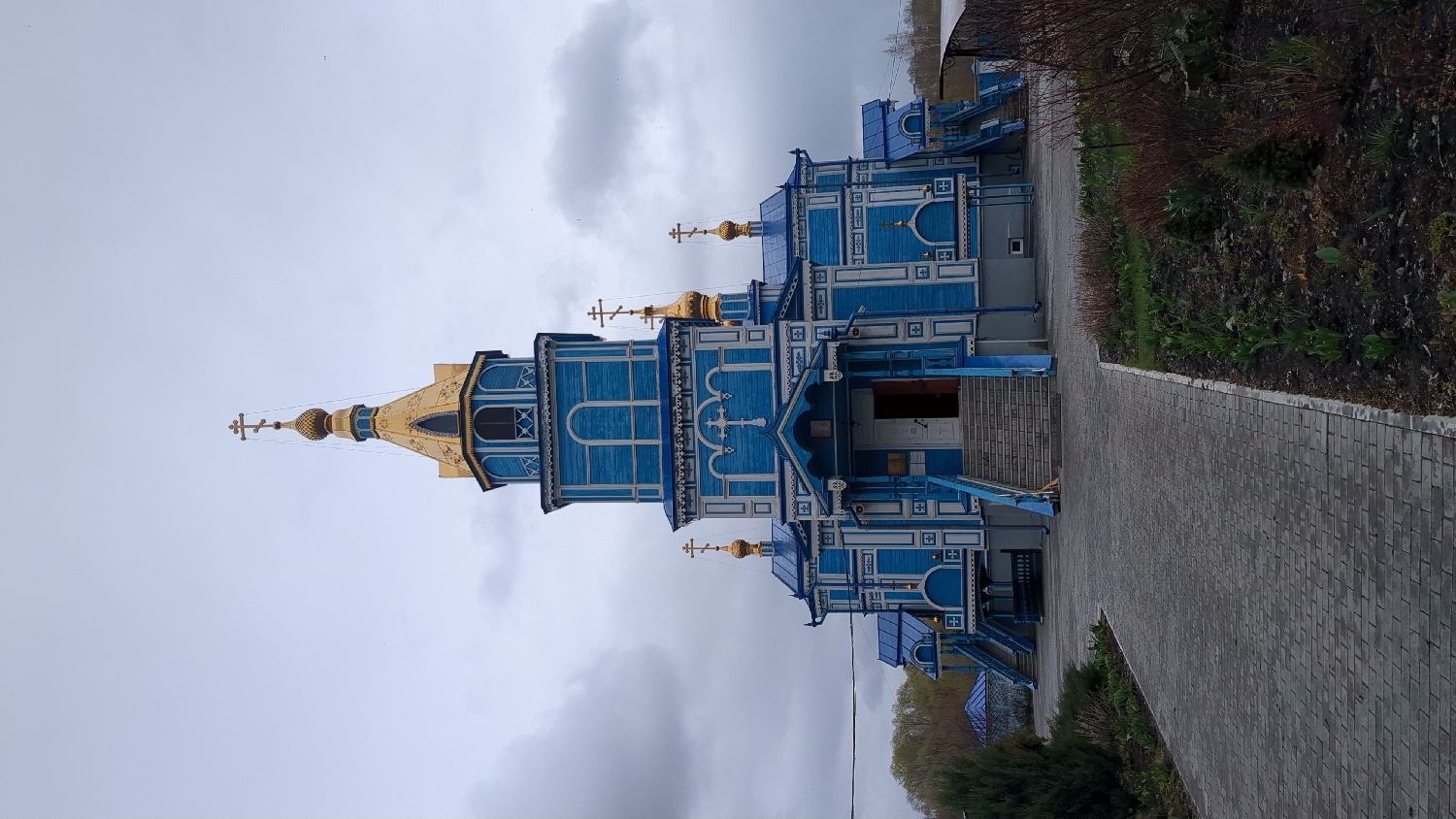 The image depicts a blue and white wooden church with golden domes and crosses, located in Ulyanovsk, Russia. The church is situated next to a paved walkway. Overcast skies fill the background. There are trees and some vegetation visible on the right.
