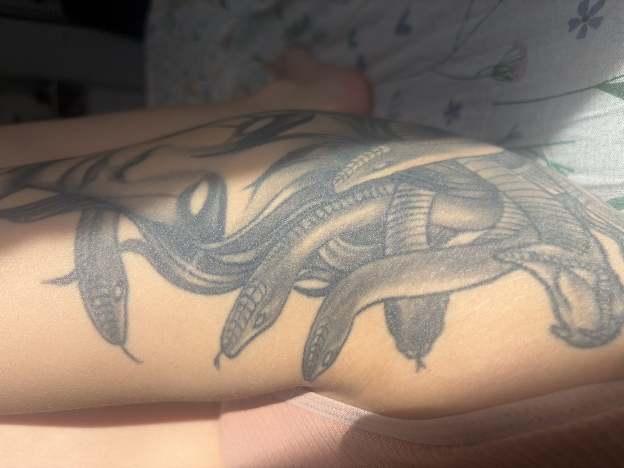 The image shows a close-up of a person's arm with a tattoo depicting Medusa's head with snakes. The arm is light-skinned. Part of a pink garment is visible at the bottom of the frame. In the background, there is a foot and a floral-patterned sheet. The location is not in Saint Petersburg, Russia.