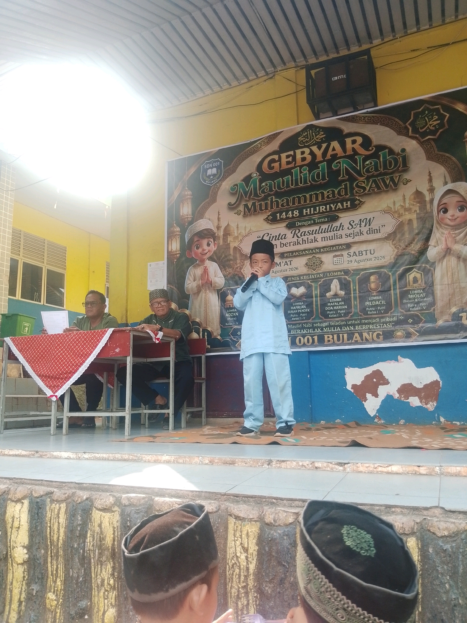 This is a photograph taken outdoors, likely in front of a building. A large banner hangs in the background, celebrating "GEBYAR MAULID NABI MUHAMMAD SAW 1448 HIJRIYAH." Below the title, it mentions the theme "Cinta Rasulullah SAW & berakhlak mulia sejak dini" (Love Prophet Muhammad SAW & have noble character from a young age). Further details on the banner indicate that the event includes various competitions like reciting the Quran, prayers, and "Pildacil" (children's Islamic lectures), and it is scheduled for Saturday, August 29, 2026.
In the foreground, a young boy dressed in a light blue outfit and a black peci (cap) stands on a stage, holding a microphone and speaking. To his left, two men are seated at a table. The man on the left, wearing a green shirt and glasses, is looking at a paper. The man on the right, also in a green shirt and peci, is looking towards the boy. A red and white checkered tablecloth covers part of the table.
In the lower portion of the image, two more young boys, viewed from the back, are also wearing pecis. They appear to be part of the audience. The background features a yellow wall and a window on the left, and a blue wall with some colorful artwork on the right. The overall setting suggests an event related to a school or community gathering celebrating Islamic heritage. The presence of an "AMT" logo on a speaker mounted on the ceiling suggests the event is indoors, or partially covered by a roof structure. The architectural details and general ambiance do not specifically point to Batam, Indonesia, but the banner's content strongly indicates an Islamic cultural event within Indonesia.
