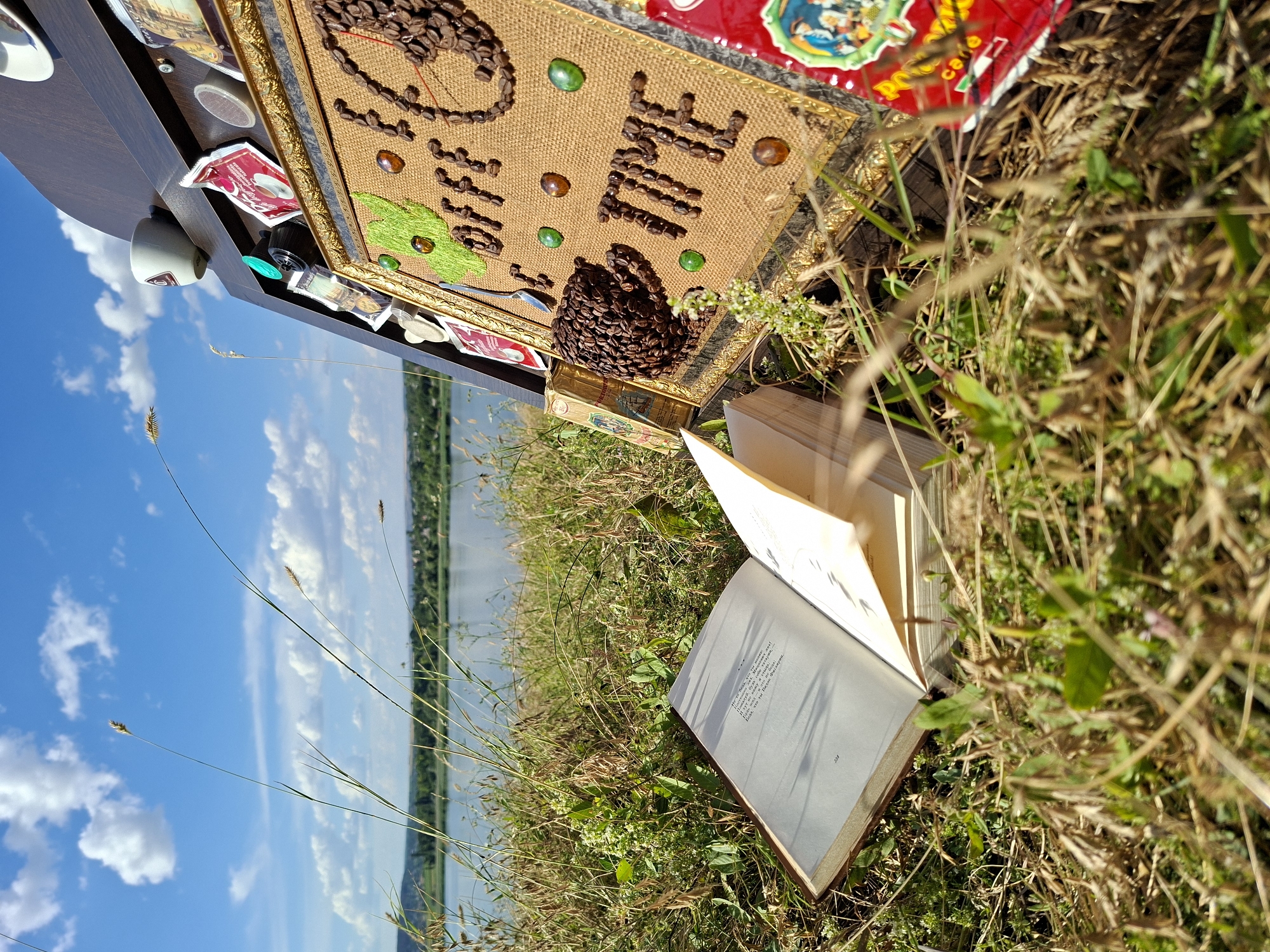 In Oxentea, Moldova, a composition is set against a backdrop of a tranquil lake and a bright, cloudy sky. An open book lies on the grassy shore, its pages filled with text. To the right, a dark wooden easel displays a canvas decorated with coffee beans, forming the words "COFFEE TIME" and a coffee cup motif. Various coffee-related items, including coffee capsules and small packets, are attached to the easel. The scene is bathed in natural light, suggesting an outdoor daytime setting.