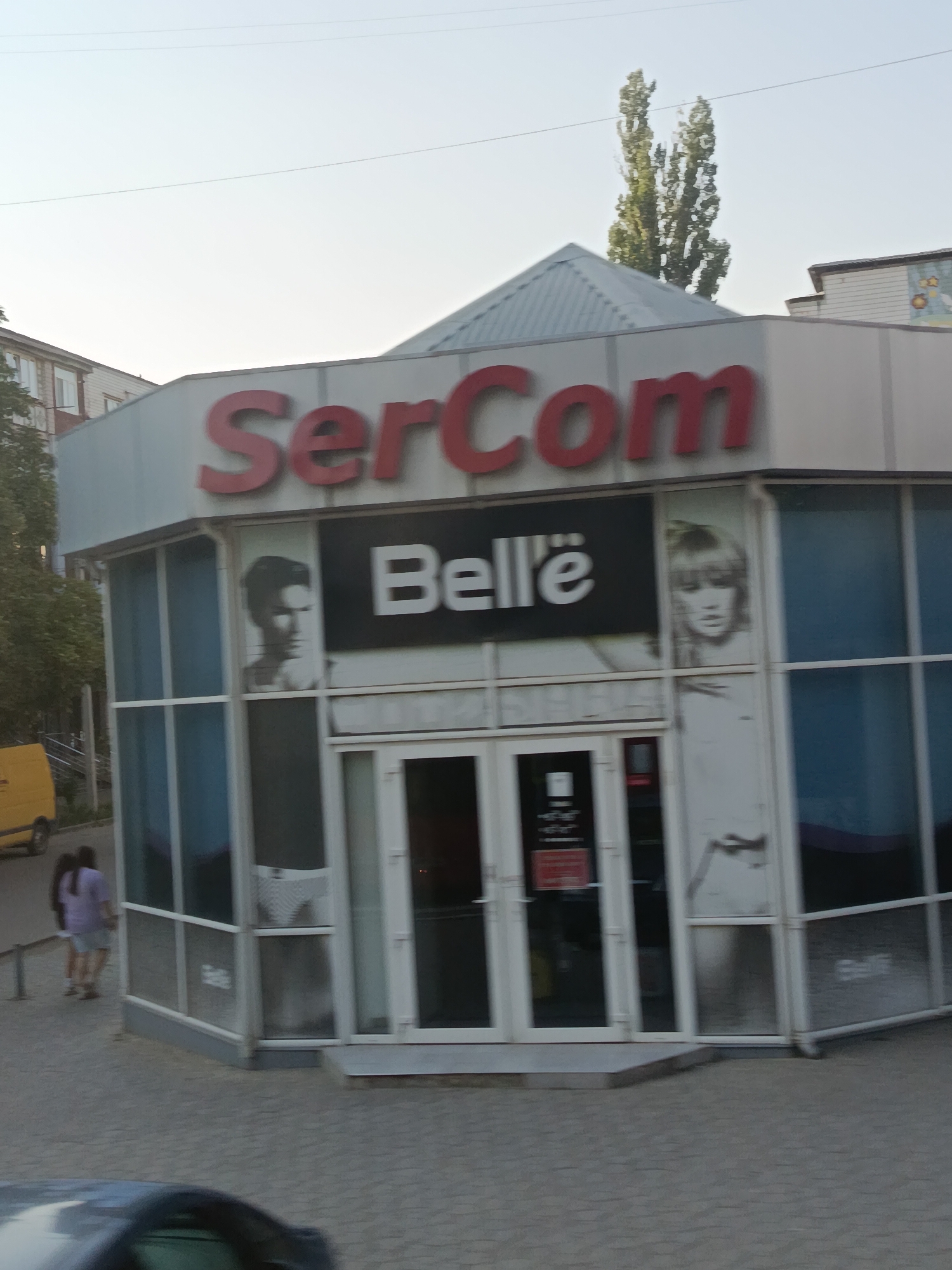 The image shows a street-level view of a commercial building with a prominent "SerCom" sign in large red letters. The building has a modern, angular design with large glass windows and a faceted roof. On the windows, there are images of a man's profile and a woman's face, suggesting the business might be a salon or a store related to beauty or personal care. Below the "SerCom" sign, another sign reads "Belle" in white letters on a black background, further reinforcing this idea.
In front of the building, there are two women walking away from the camera. One is wearing a purple top and shorts, and the other is wearing a light-colored top and shorts. They are walking on a paved sidewalk. To the left of the building, a yellow van is parked. The overall scene suggests an urban or suburban commercial area.
The lighting indicates it is either early morning or late afternoon, with the sun casting long shadows and the sky appearing bright. The weather seems to be clear and calm. The presence of the "SerCom" and "Belle" signs, along with the imagery on the windows, provides specific textual information about the businesses operating here. The setting appears to be a business district or a shopping area in Cahul, Moldova. The people are casually dressed, and there are no obvious signs of any particular event or strong emotions. The focus is on the storefront and its signage, highlighting the commercial nature of the location.