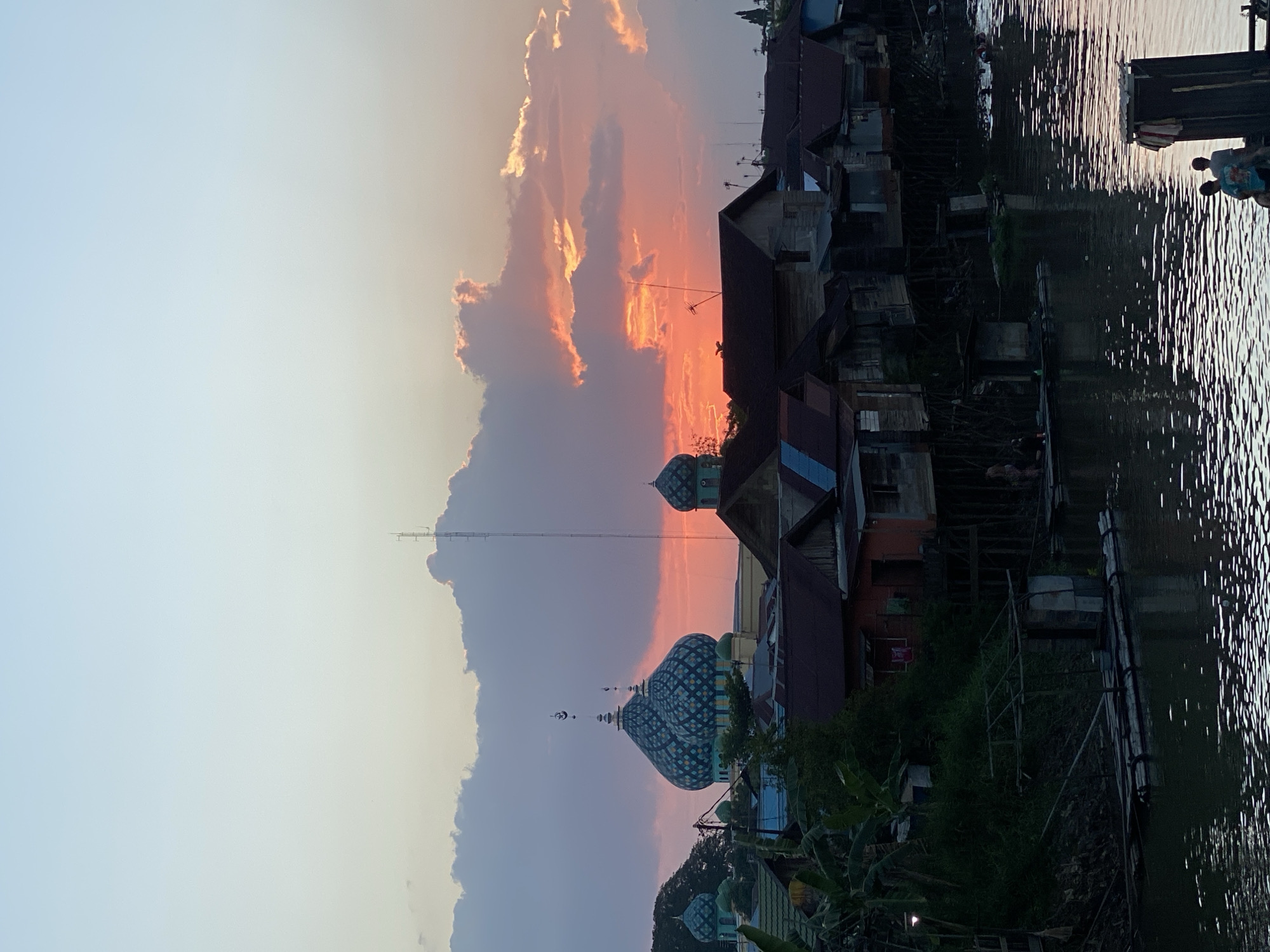 The image depicts a sunset scene in Tunggul Irang Ulu, Indonesia, with a focus on a riverside settlement and a prominent mosque. The sky is filled with colorful clouds, ranging from soft pastels to vibrant oranges and reds, illuminated by the setting sun.
In the foreground, a body of water reflects the colors of the sky. Along the bank of the river, a row of stilted houses is visible, constructed from wood and corrugated metal, showing signs of wear. Some of the houses have roofs with distinct blue and red sections. A small raft or wooden platform is moored on the water near the houses, and a few people are standing on it. Further along the river, more buildings and structures can be seen.
Dominating the skyline are the ornate domes of a mosque, characterized by their intricate blue and white geometric patterns. A tall, slender tower or antenna stands behind the mosque. Lush green vegetation and trees are interspersed with the buildings, adding a natural element to the urban landscape. The overall impression is one of a tranquil evening in a riverside community, where traditional architecture meets the natural beauty of the sunset.