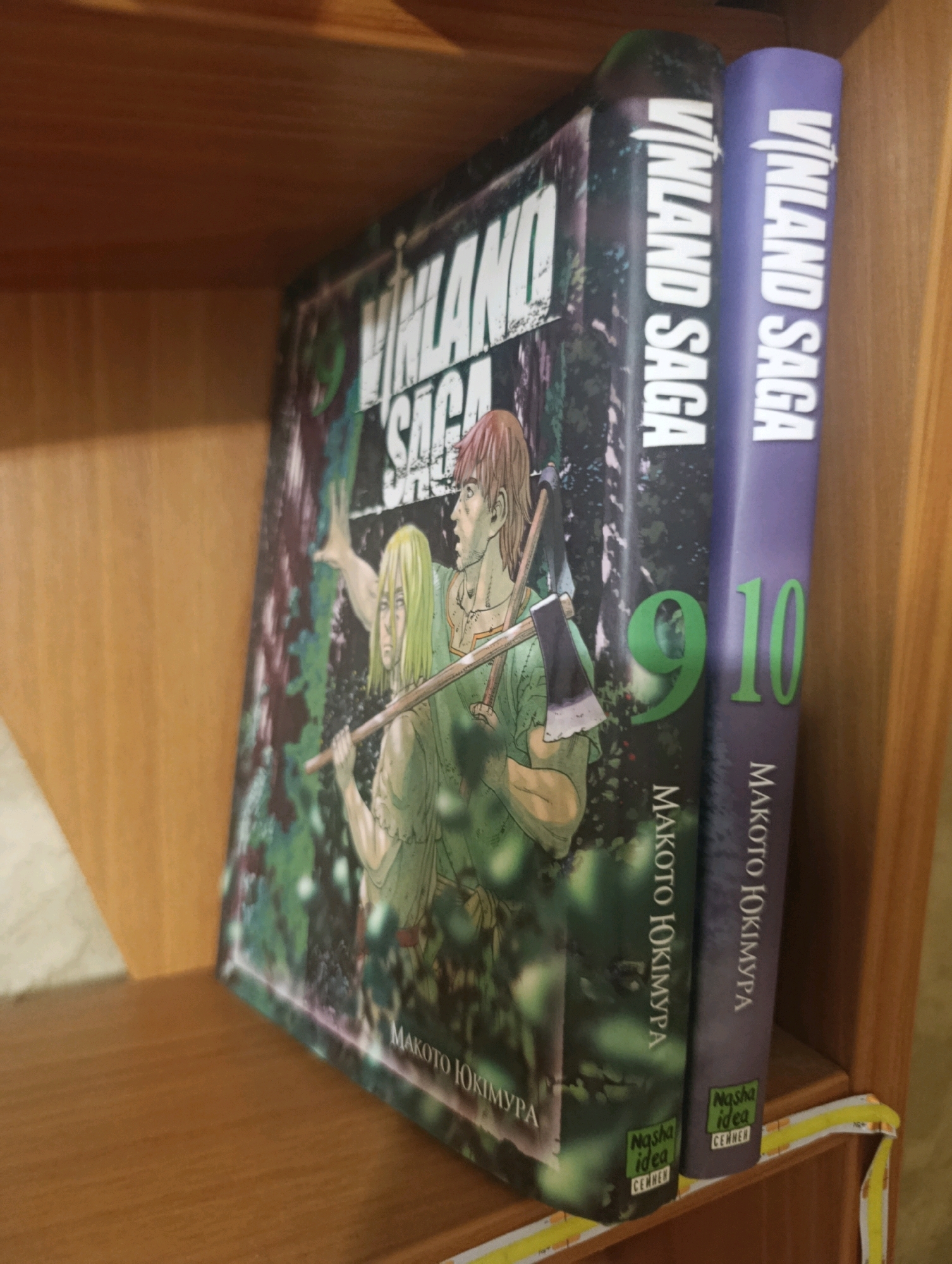 The image displays two manga volumes of "Vinland Saga" placed on a wooden shelf. The left volume has a cover illustration of two characters in a forest setting. The right volume is solid purple with "Vinland Saga" printed vertically. Both volumes have "Makoto Yukimura" printed vertically and a "Nasha Idea" logo at the bottom. The number "9" is visible on the left volume and "10" on the right. The lighting suggests an indoor environment, possibly a home or bookstore. There is no indication of the specific location within Horishni Plavni, Ukraine.
