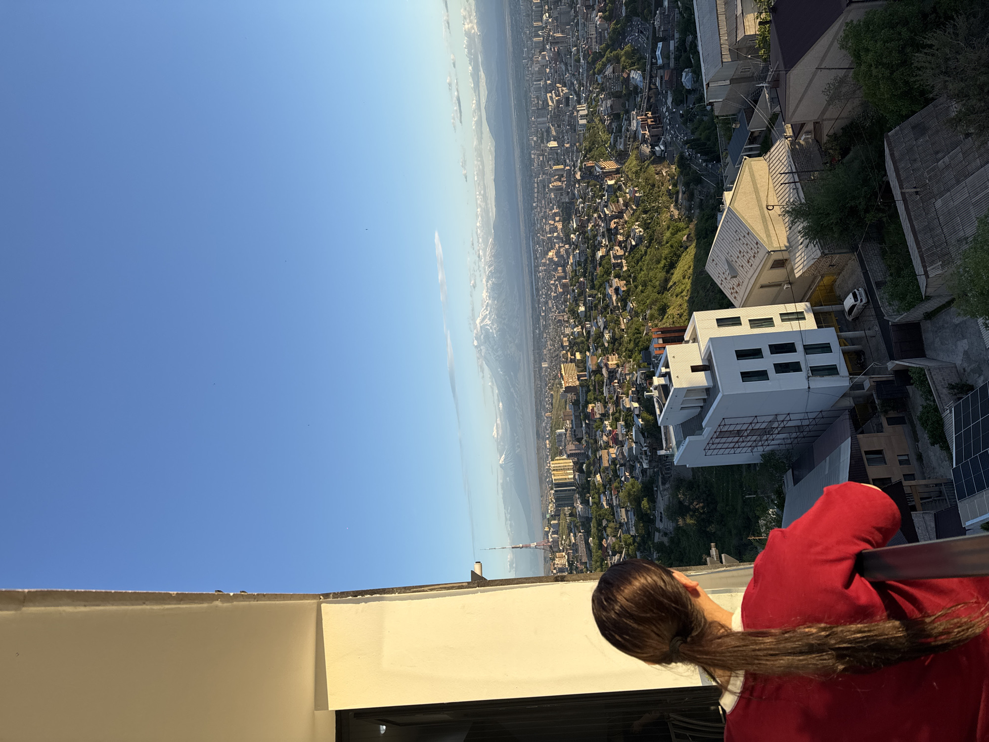 A medium shot taken from a high vantage point overlooks a city. A person with long brown hair, wearing a red shirt, leans against a railing, appearing to look out over the scene. In the distance, a sprawling urban landscape is visible, featuring a mix of buildings, trees, and infrastructure. A television tower is visible above the buildings, identifying the city as Yerevan, Armenia. The sky overhead is a clear blue, with a hint of clouds on the horizon. The scene appears to be captured during the daytime.