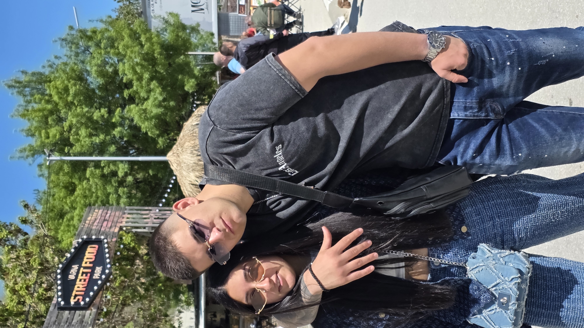 The image shows a couple posing for a picture in what appears to be a street food park in Varna, Bulgaria. The man is wearing a dark gray T-shirt, blue jeans with white paint splatters, and a silver wristwatch. He has a black shoulder bag. The woman has long, dark hair, and is wearing a denim jacket and a small blue purse with a chain strap. Both are wearing sunglasses. In the background, there is a sign that says "Varna Street Food Park" and other people sitting at tables. There is a straw-covered structure and green trees visible. The sky is blue. A sign that includes "ПЛАЖ" (beach) in Cyrillic is also visible, potentially indicating proximity to a beach resort.