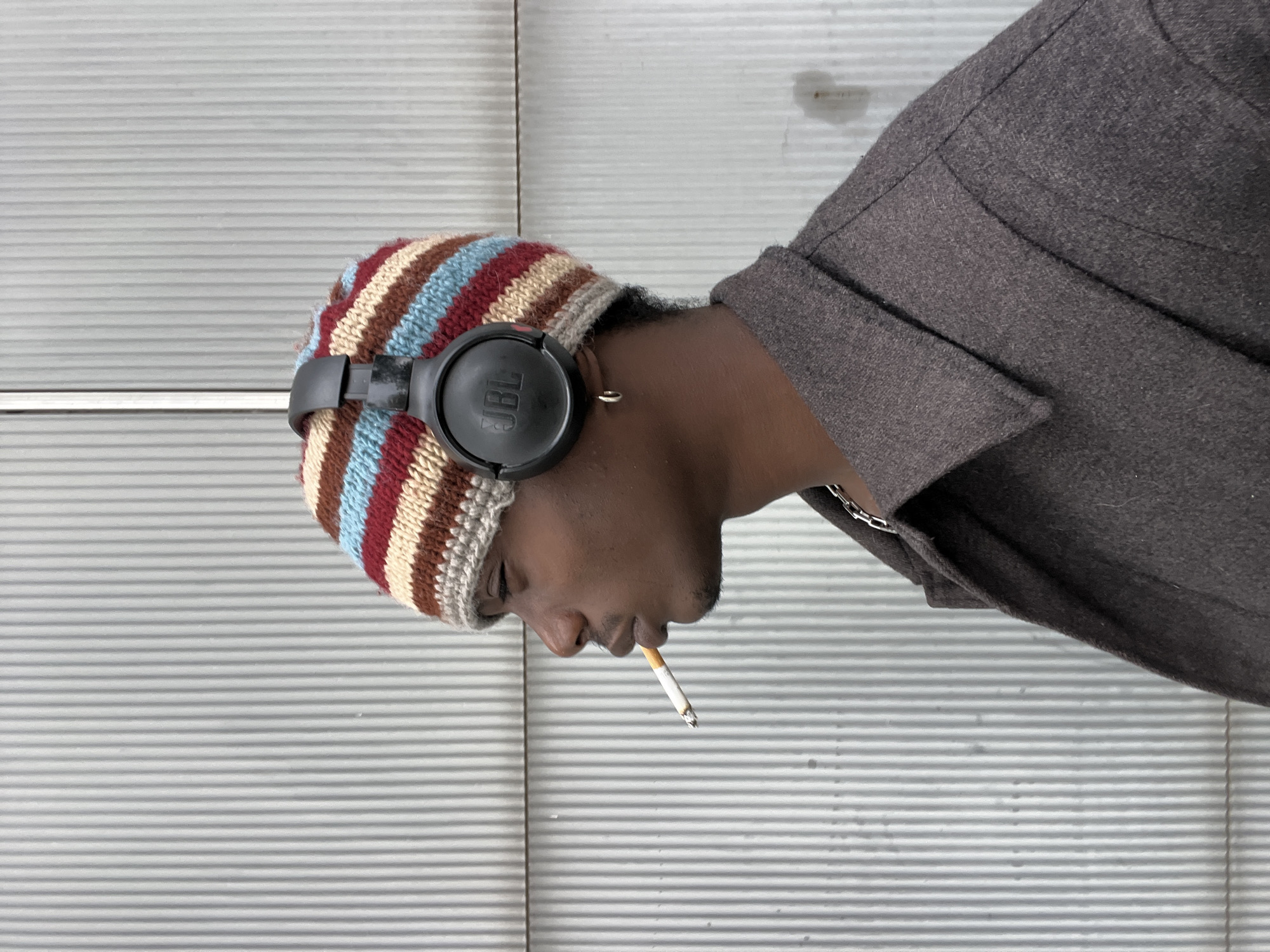 The image features a person, likely a young adult male with dark skin, shown in profile from the chest up. They are positioned on the right side of the frame, with their head slightly bowed and facing towards the left. The person wears a multi-colored striped knit beanie with hues of red, blue, beige, and brown. Black JBL over-ear headphones are worn over the beanie, with the "JBL" brand name visible on the left earcup. A lit cigarette, showing a glowing ember, is held between their lips. A silver hoop earring is visible in their right ear, and a silver chain is partially seen around their neck, covered by a dark grey or brown jacket/coat with a visible collar. The person's eyes appear closed or cast downwards, conveying a pensive or relaxed expression.
The background is a light grey wall with distinct, uniform vertical stripes, suggesting a corrugated metal surface or a roll-up door. A few faint smudges or stains are discernible on the wall. The overall setting implies an urban or industrial environment. No specific time of day or weather conditions are indicated, with diffused lighting present. While the location context is provided as Barcelona, Spain, the image itself contains no specific visual cues to identify this city.