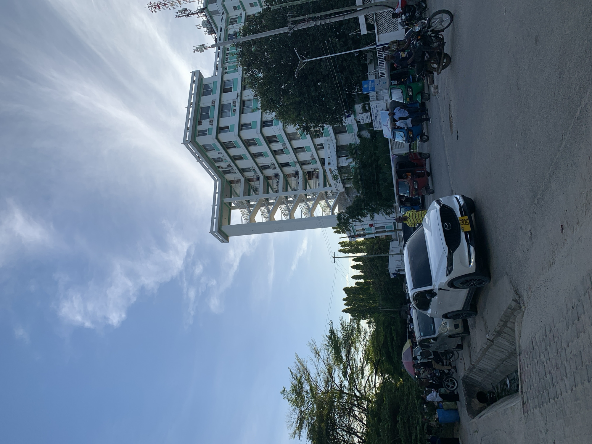 A street scene in Dar es-Salaam, Tanzania. A modern multi-story building with a white exterior and turquoise accents is prominently featured. An elevated support structure is connected to the building. Utility poles and wires are visible along the street. Parked vehicles, including a white SUV with a visible license plate, tuk-tuks and motorbikes line the street. Trees provide foliage in the scene. The sky is blue with wispy clouds.