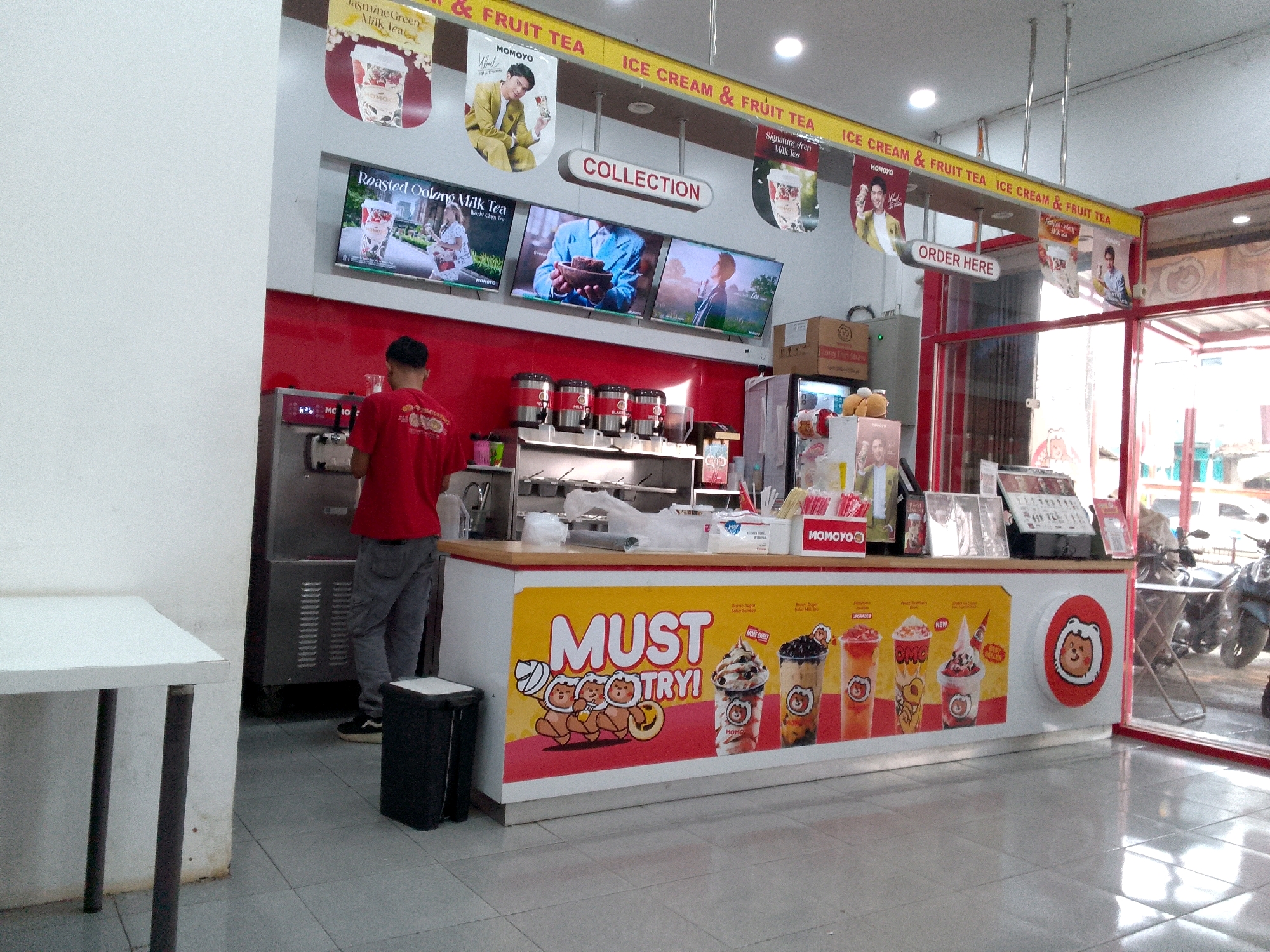 The image shows the interior of a Momoyo store, a bubble tea and ice cream shop located in Rajamandala Kulon, Indonesia. A male employee in a red uniform is standing behind the counter, preparing drinks. The counter is decorated with images of various beverages and ice cream, with the slogan "MUST TRY!" prominently displayed. Above the counter, several screens advertise different menu items, including "Roasted Oolong Milk Tea." A trash can is positioned near the counter. In the background, a refrigerator and an ice cream machine are visible. The store appears to be clean and well-lit, with a red and white color scheme predominating. Outside the store, motorcycles can be seen parked.