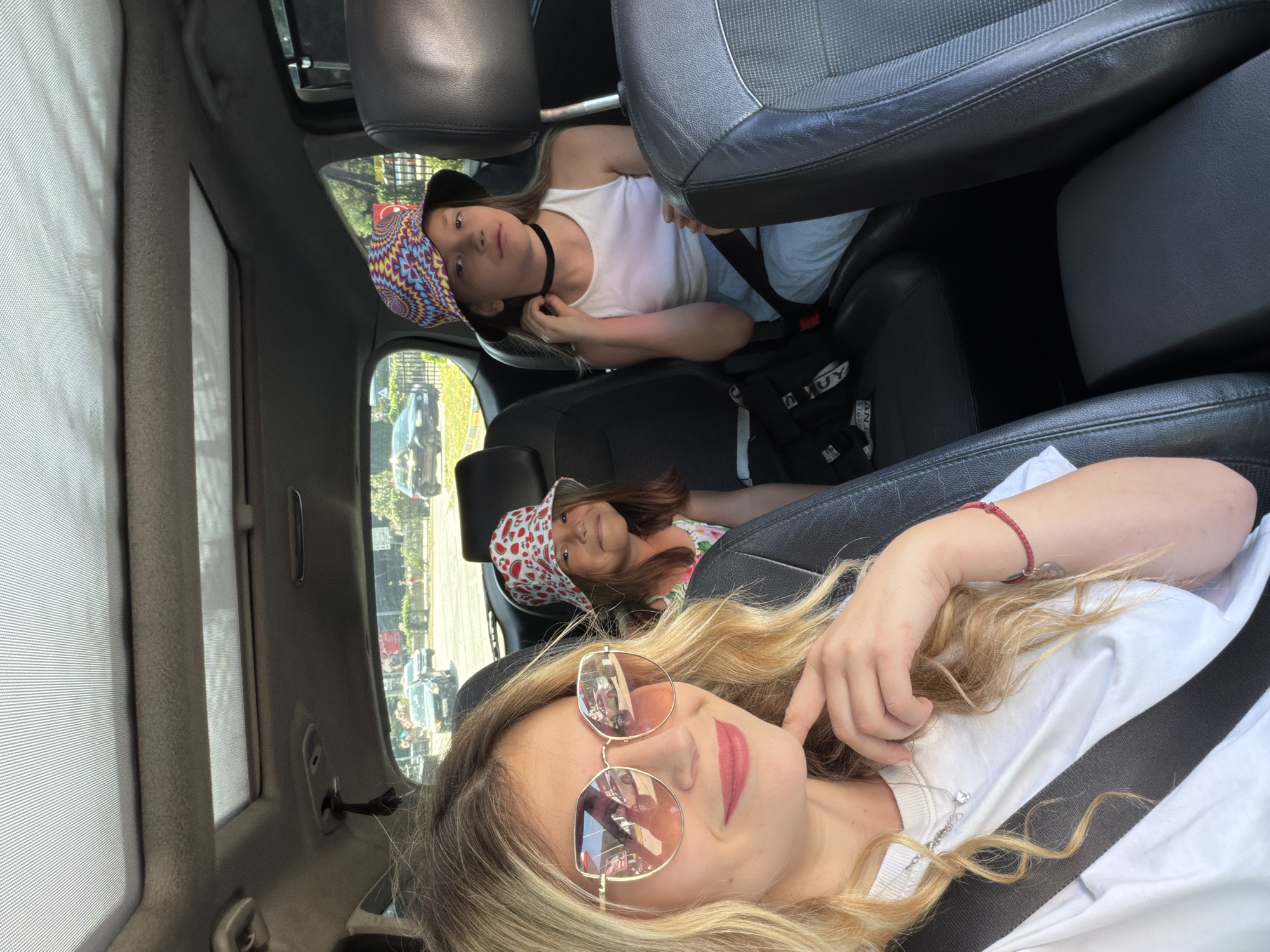 An interior vehicle photograph depicts an adult female and two children within what appears to be a multi-passenger car on a sunny day in Budva, Montenegro.
In the foreground, an adult female with long blonde, wavy hair is seated in the front passenger seat, wearing a white t-shirt and a fastened seatbelt. She wears large, gold-rimmed sunglasses with a pink gradient tint, reflecting the street scene outside. Her left hand, adorned with a red string bracelet, is positioned near her chin. A silver chain necklace with a heart pendant is visible. She smiles faintly, looking directly towards the camera, suggesting a selfie.
Behind the adult female, in the left rear seat, a younger child with brown hair is partially visible. She wears a white bucket hat patterned with red watermelons and a light-colored top with a floral or similar pattern. This child is smiling and looking towards the camera, also secured by a seatbelt.
To the right, in the rear passenger seat, an older child with long brown hair is seated. She wears a multi-colored bucket hat with a geometric pattern, a white sleeveless top, light blue pants, and a black choker necklace. Her right hand is raised to her neck near the choker. She maintains a neutral expression, looking towards the camera. A black bag with white lettering is visible on the seat next to her. She is also secured by a seatbelt.
The vehicle's interior features black leather front and middle seats, with a darker grey fabric for the rear right seat. The roofliner is grey, and two panoramic sunroofs with perforated covers are visible overhead. A dark grey center console is present between the front seats.
Through the vehicle's windows, a city street scene is visible. Other vehicles, including a dark sedan, are present on an asphalt road. Green foliage, a black metal fence, and indistinct buildings are observable in the background under a clear, bright sky. A red sign is partially visible on the right side of the background. The lighting suggests midday or early afternoon conditions.