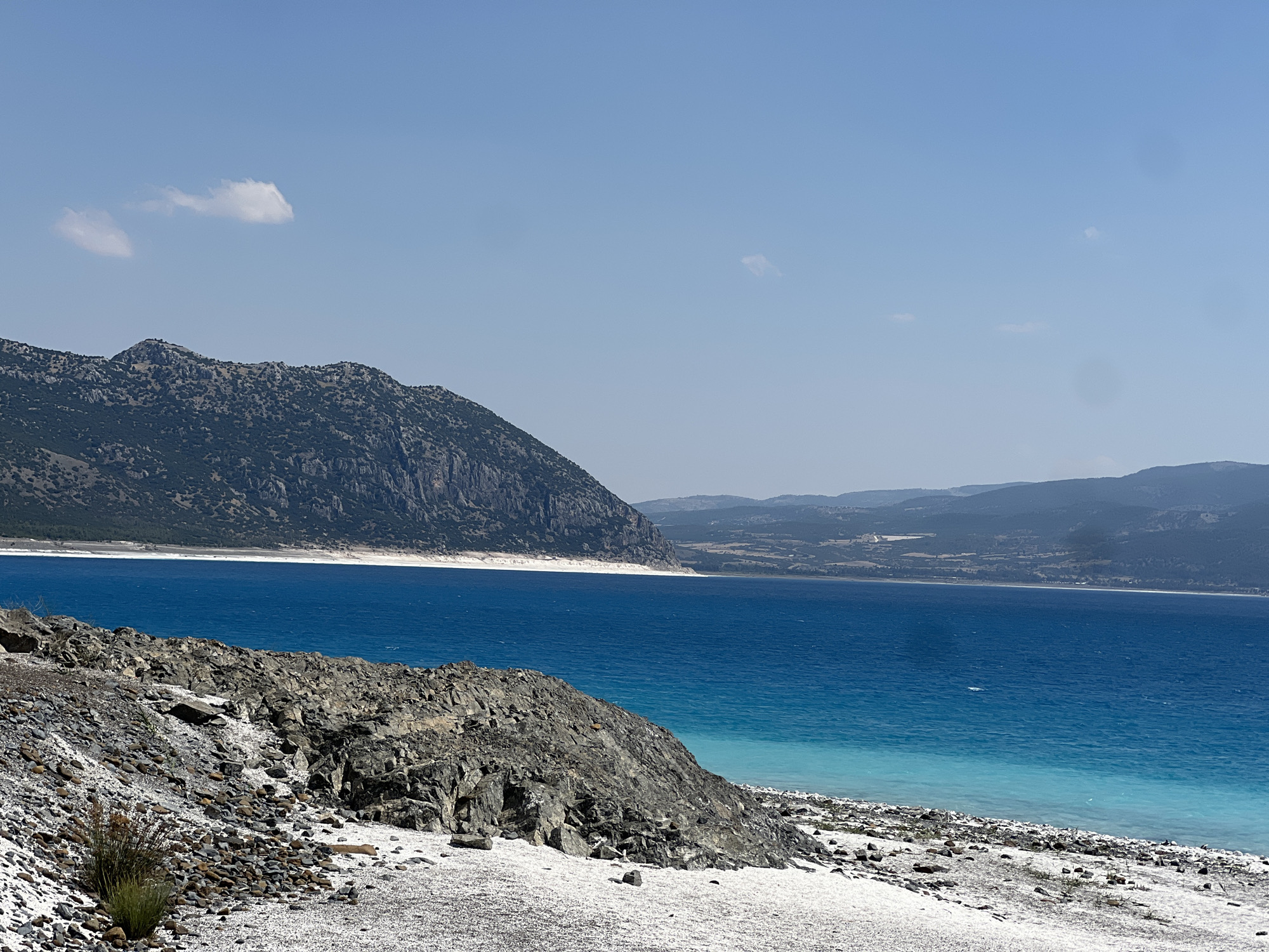 The image depicts a scenic view of Salda Lake in Yeşilova, Turkey. The foreground is dominated by a rocky, white-sand shoreline with scattered pebbles and sparse vegetation. A large, dark grey rock formation juts out into the frame from the left. The middle ground features the strikingly turquoise and deep blue waters of Salda Lake, with a distinct transition in color indicating depth. In the background, a vast, mountainous landscape covered in vegetation stretches across the horizon, meeting a clear, pale blue sky with a few wispy clouds. The environment is natural and tranquil, with no people or man-made structures visible in the immediate scene.