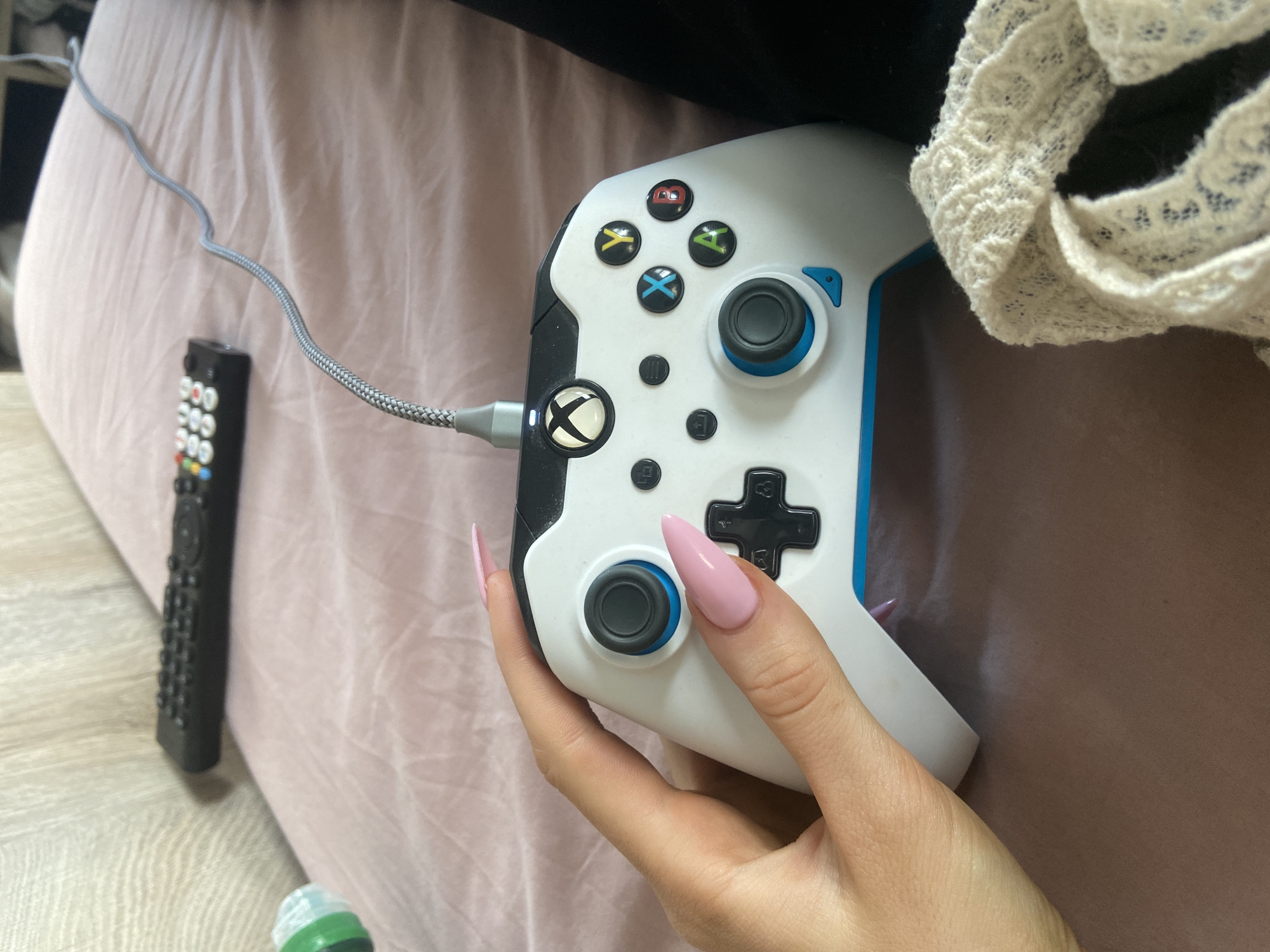 The image shows a person holding an Xbox controller. The controller is white with blue accents and is connected to a cable. A television remote lies next to it on a pink sheet. The scene is indoors. No information suggests a London, United Kingdom location.