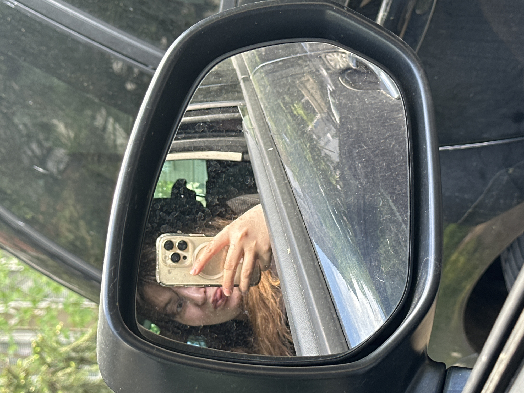 The image shows a selfie reflected in a car's side mirror. The reflection includes the face of a person with long brown hair, holding a smartphone with multiple camera lenses, presumably taking the photograph. The mirror is black and rectangular with rounded corners. In the background, visible through the mirror and around its edges, is a blurred cityscape with greenery. The overall scene seems to be outdoors, during the day. There is no information in the image to confirm a location.