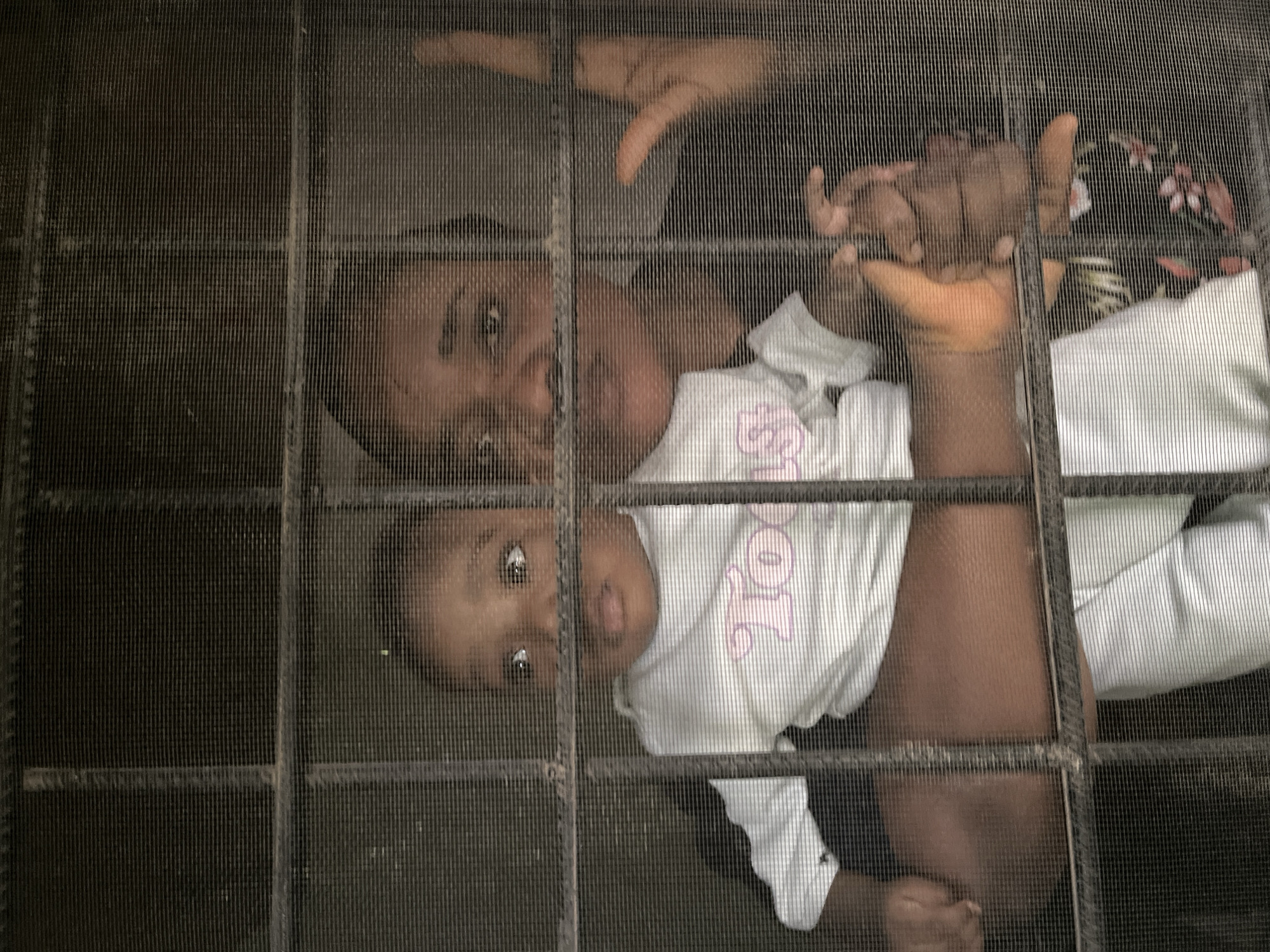 The image, taken in Barakin Akawo, Nigeria, shows two people, an adult and a child, looking through a window screen. The adult, partially visible in the top half of the frame, has their hands raised, with one hand resting on the screen and the other reaching out. They appear to be looking downwards. The child is positioned below the adult, facing the camera. They are wearing a white t-shirt with a pink graphic. The child's eyes are open, and their mouth is slightly parted, suggesting they are looking at something or someone. The background is dark and indistinct, with hints of floral patterns on clothing worn by the adult. The scene is viewed through the grid of the window screen, which creates a textured overlay on the image. The lighting suggests it might be daytime, but the overall mood is subdued. No specific emotions are clearly discernible due to the distance and the screen. There is no visible text, aside from the graphic on the child's shirt.