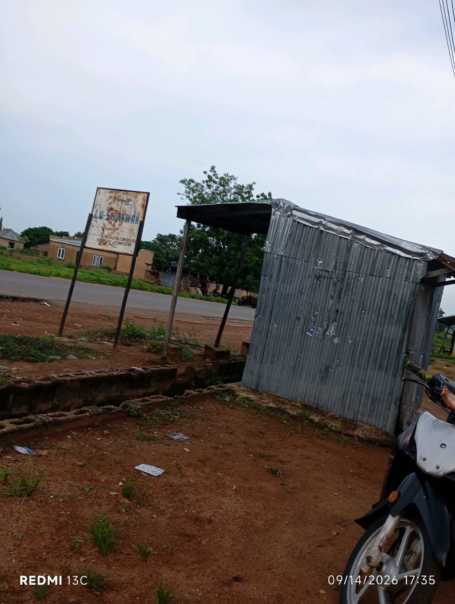 The image depicts a roadside scene in Shendam, Nigeria. In the foreground, the ground is covered in reddish-brown dirt with scattered green grass and some white debris, possibly litter. A concrete drainage channel runs along the left side of the frame. Behind this, a weathered signpost with the inscription "LU-SHANWAN" is visible, propped up by two metal poles.
Further back, a street runs horizontally across the image, with a few buildings and trees lining its opposite side. To the right of the signpost is a small, corrugated metal structure, appearing to be a makeshift shelter or stall, with a sloped roof. A portion of a black and white motorcycle is visible in the lower right corner of the image, angled towards the viewer. The sky is overcast with a pale, cloudy appearance. The overall impression is one of a rural or semi-urban setting in Nigeria.