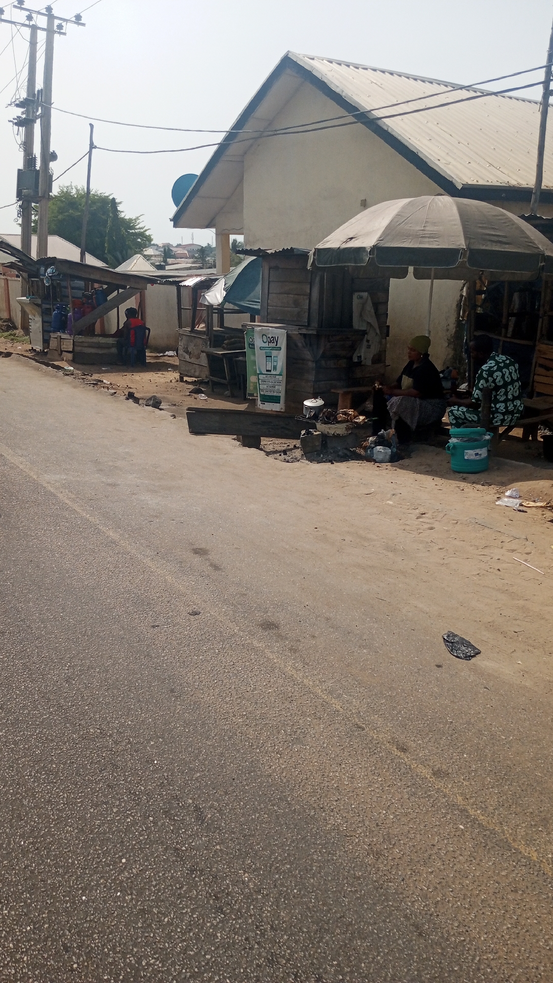 This street scene, set in Lokoja, Nigeria, captures a snapshot of daily life along a commercial road under bright, clear skies. The foreground is dominated by an asphalt road, partially marked with a faded yellow line, which gives way to a dirt shoulder on the right.
Along this dirt path, several rudimentary wooden stalls and kiosks are visible. One prominent stall is shaded by a large, light-colored umbrella, beneath which two individuals are seated. One woman, wearing a dark top and patterned skirt, is actively engaged in what appears to be cooking or food preparation, attending to a pot over a small, ground-level fire or heat source. Beside her, another person, dressed in a patterned garment, sits partially obscured. A bright green plastic bucket rests on the ground nearby. Further down the street, a third person in red and dark attire is seated on a chair next to another stall.
A notable object in the mid-ground is a white rectangular sign displaying the "Opay" logo and the text "The Better Way to Pay," along with other smaller service-related details, indicating a mobile money or payment service point.
In the background, a light-colored building with a distinctive corrugated metal gable roof stands out, surrounded by other smaller structures and some trees, suggesting a residential or mixed-use area. On the left side of the road, tall utility poles with a network of wires and electrical components are clearly visible, signifying the area's infrastructure. The overall impression is one of informal commerce and everyday activity in a semi-urban or rural setting during daylight hours.
