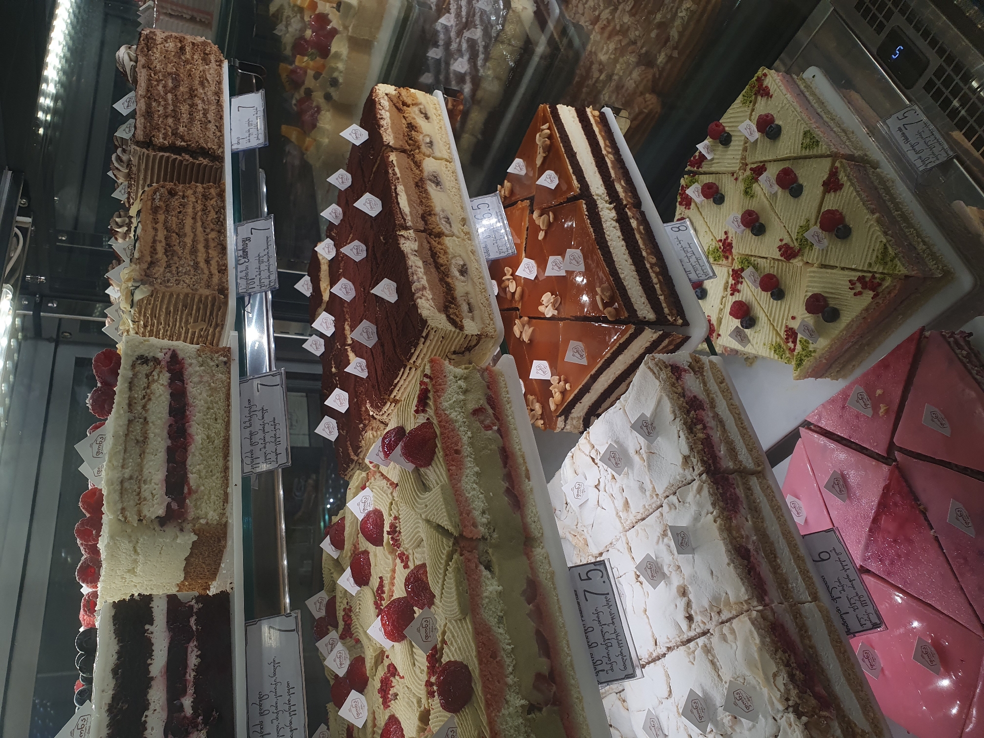 This image shows a display case filled with a variety of cakes and pastries, likely at a bakery or patisserie in Batumi, Georgia. The selection includes layered cakes with fruit and cream fillings, chocolate cakes with caramel frosting, and lighter sponge cakes. Several items have decorative elements like fresh berries, nuts, and intricate frosting designs. Small flags with a logo are placed on many of the desserts. Prices are visible on some of the labels, suggesting a retail environment. The lighting highlights the textures and colors of the baked goods, creating an appealing presentation.
