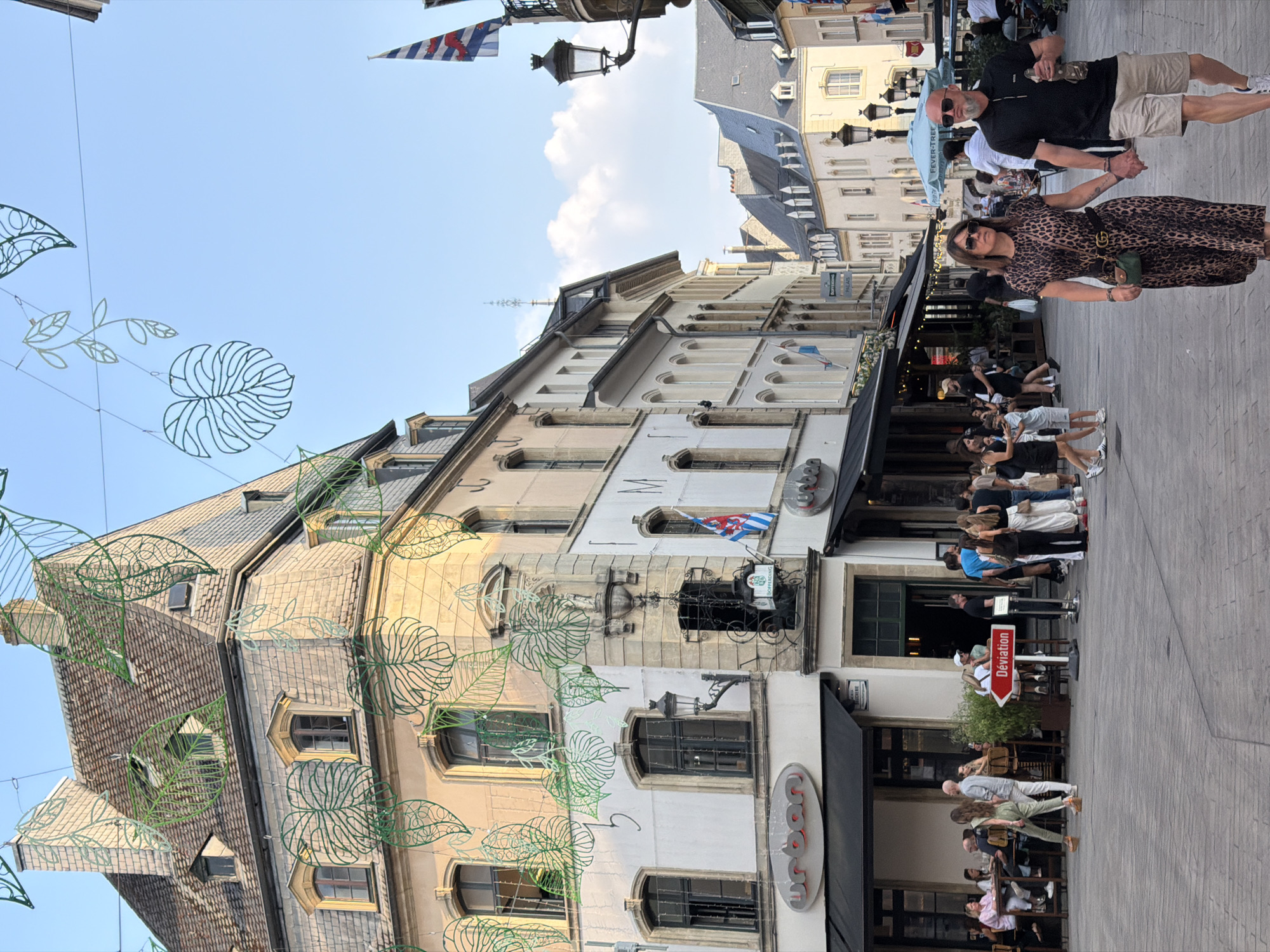 The image depicts a bustling street scene in Luxembourg City, Luxembourg. In the foreground, a couple walks hand-in-hand, the woman wearing a leopard-print dress and the man in a black polo shirt and beige shorts. Behind them, other pedestrians are visible, as are outdoor seating areas for cafes and restaurants. The buildings lining the street feature traditional architecture with stone facades and mansard roofs. Green leaf-shaped decorations are strung across the street, suggesting a festive or seasonal event. A red sign with the word "Déviation" (Detour) points to the left, indicating a potential road closure or rerouting. The sky is clear blue with a few wispy clouds. The overall atmosphere appears lively and pleasant, with people enjoying the urban environment.