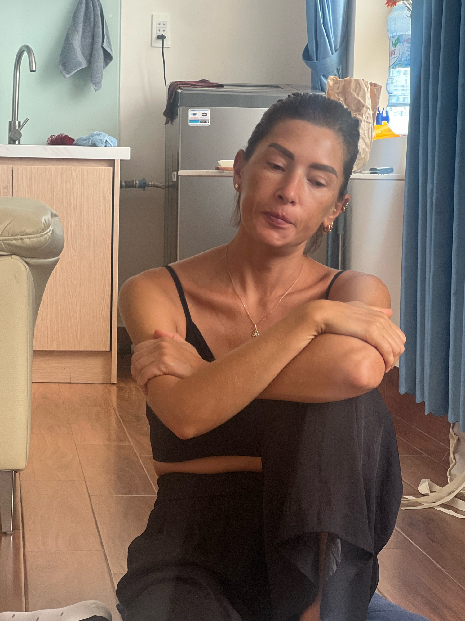 A woman sits on the wooden floor in a room with a light green wall, a wooden cabinet, a sink, a refrigerator, and blue curtains. She is wearing a black sleeveless top and black pants. Her arms are crossed over her chest, and she is looking down. A bottle of water and a brown paper bag are on a shelf behind her. A light-colored sofa is to her left. The scene is consistent with an apartment in Đà Nẵng, Vietnam.