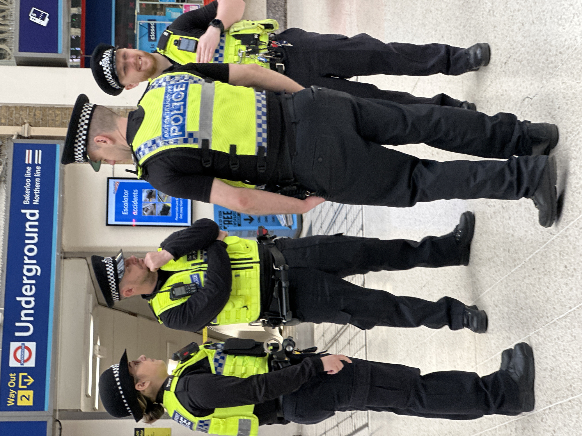 Here is a description of the image:
The image shows a group of four uniformed British Transport Police officers standing in a row inside an Underground station in London, UK. The officers are wearing black trousers, black shirts, and bright yellow vests with "POLICE" written on them. They also wear traditional black police caps with a checkered pattern. Signs in the background indicate "Underground," "Way Out 2," "Bakerloo line," and "Northern line." A screen mentions "Escalator accidents." The officers are standing on a tiled floor. The environment appears to be an area of the station with signage and possibly advertisements.