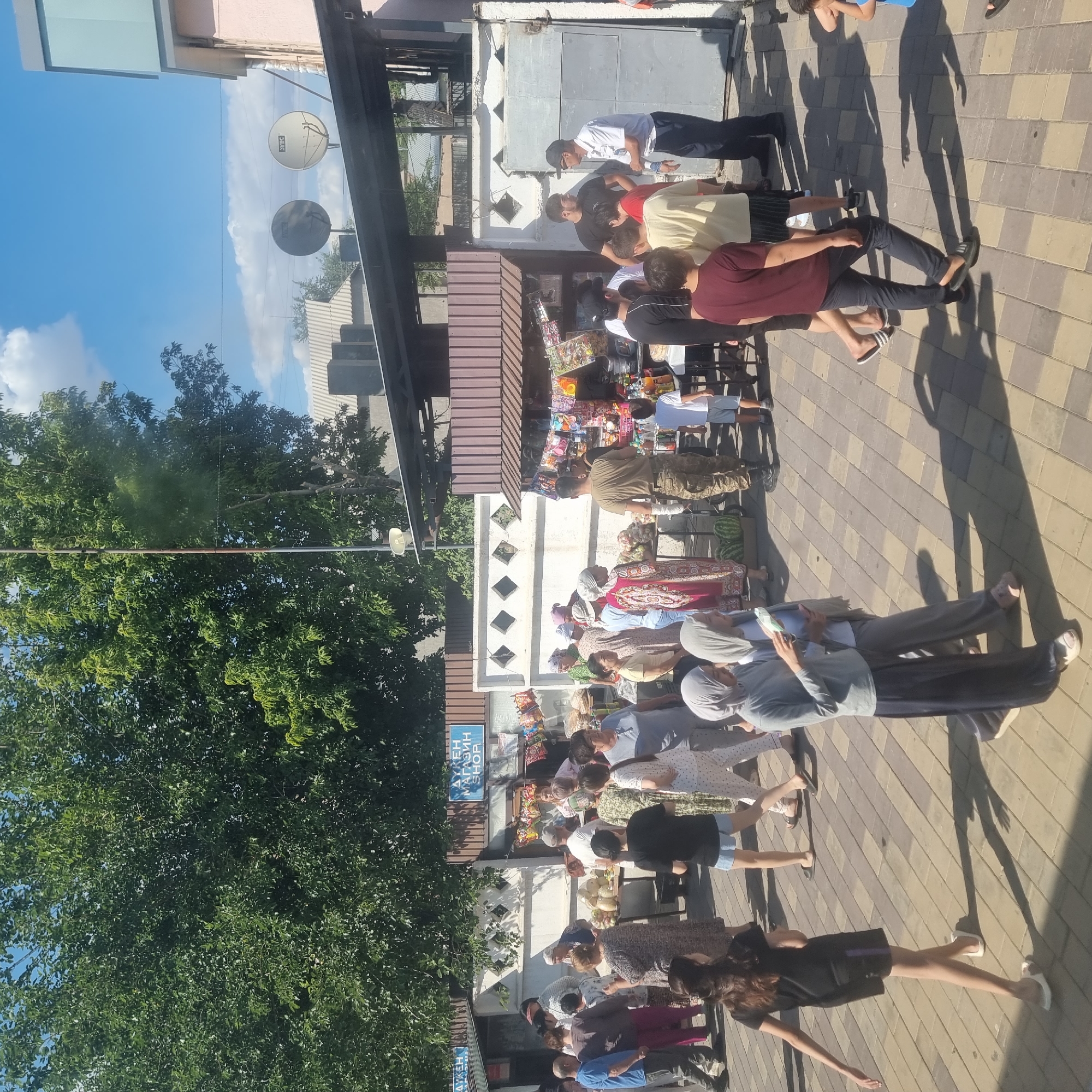 The image depicts a bustling outdoor market scene, likely in Turkistan, Kazakhstan, on a sunny day. Several stalls are set up, displaying various goods including packaged snacks, drinks, and produce such as watermelons and other fruits. A group of people are browsing the stalls and interacting with vendors. The architecture suggests a blend of modern and traditional elements, with corrugated metal roofing and simple stall structures. The ground is paved with interlocking bricks. The sky is bright blue with scattered clouds, and lush green trees are visible in the background, providing shade. The overall atmosphere is one of daily commerce and community activity.