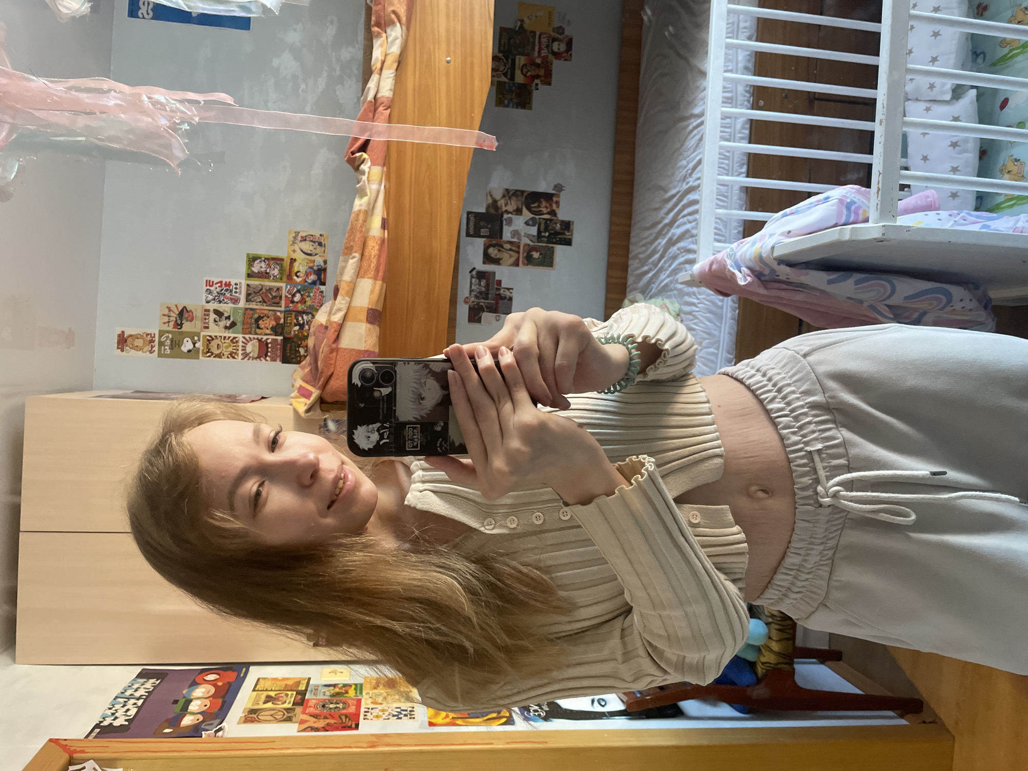 A young woman stands in front of a mirror, taking a selfie with her phone. The reflection shows a cluttered room, with a bed, wardrobe, and various items scattered around. The room features posters and decorations on the wall. The woman wears a light-colored sweater and sweatpants. No indication the location is in Siva, Russia.