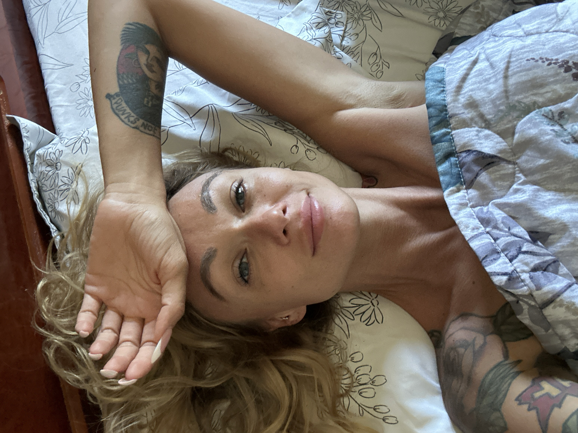 A woman with blonde hair and a tattoo on her arm lies in bed. She is holding her hand up to her forehead and looking at the camera. The bed is covered with a floral patterned duvet and sheets. The scene appears to be indoors, in a bedroom. There is no indication of the specific location in Mũi Né, Vietnam.