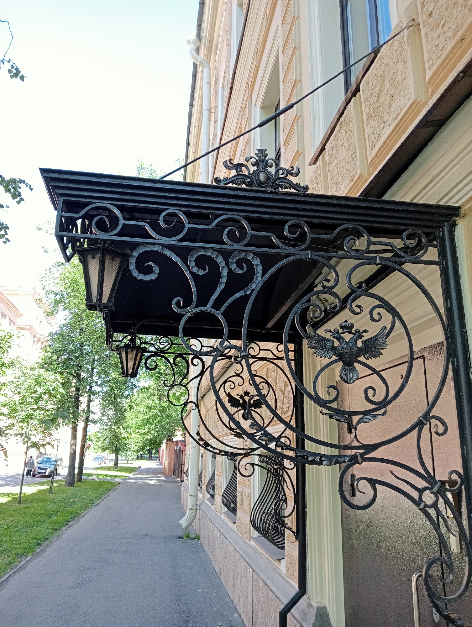 The image depicts an ornate wrought-iron canopy over the entrance to a building on a sunny day. The canopy features intricate scrollwork and decorative elements, including what appears to be a double-headed eagle motif. Two lanterns are suspended from the canopy. To the left of the entrance, a paved sidewalk runs alongside a grassy area with trees and a street. A silver SUV is parked on the street, and a person is standing next to it. Further down the street, more trees and buildings are visible. The building itself has a textured beige facade and a white drainpipe. The architectural style suggests an older European city, possibly Saint Petersburg, Russia, given the presence of a double-headed eagle, a symbol historically associated with Russia. The overall scene conveys a sense of historic urban charm.