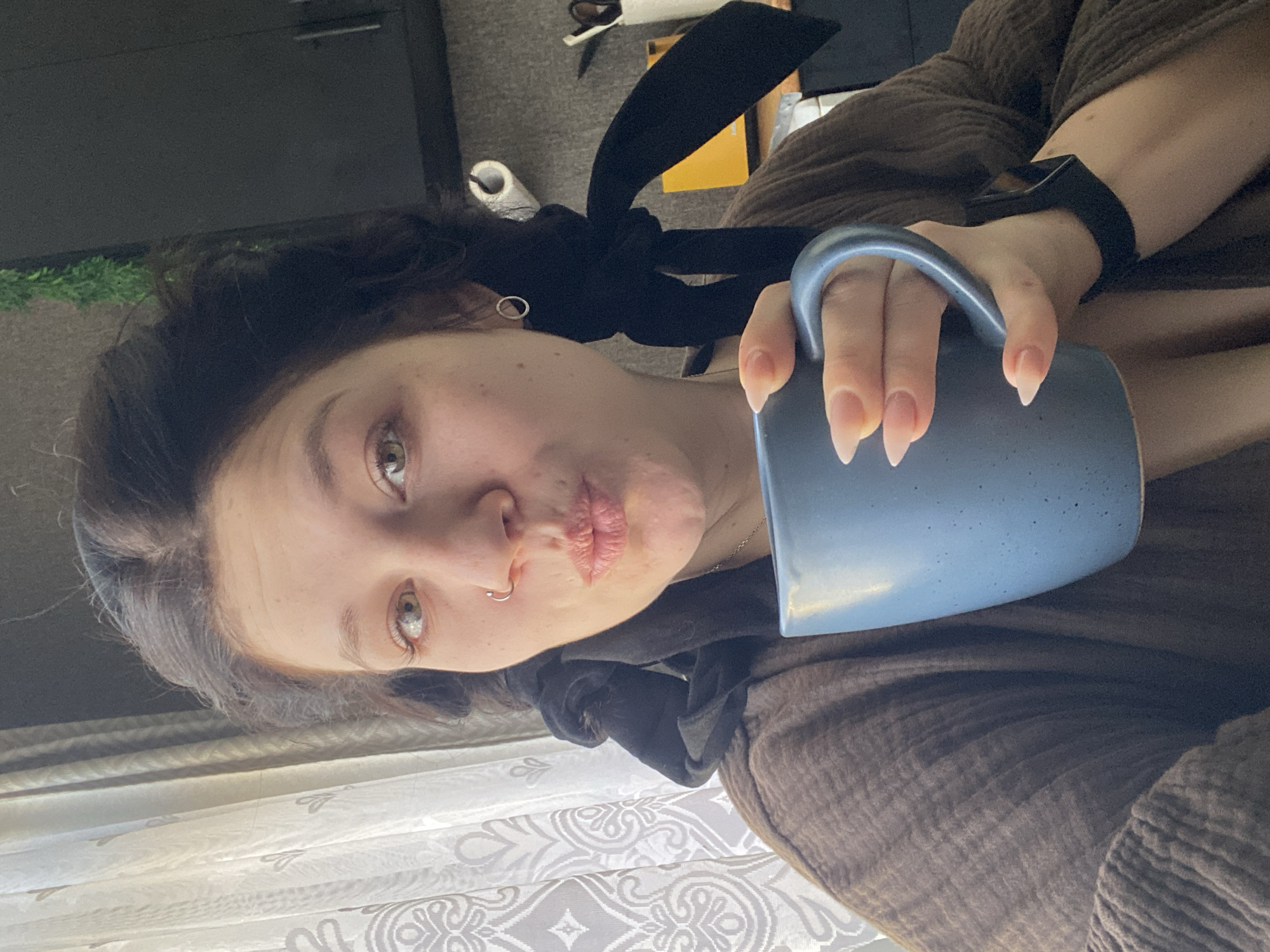 A young woman with fair skin, light eyes, and dark hair pulled back with a black bow is holding a blue speckled mug. She has a nose ring and is making a kissing face towards the camera. She is wearing a brown, textured garment. A black fitness tracker is visible on her wrist. The background includes a window with white patterned curtains, a dark cabinet, and some greenery. There is no indication that the location is in the city of Chaikovsky, Russia.