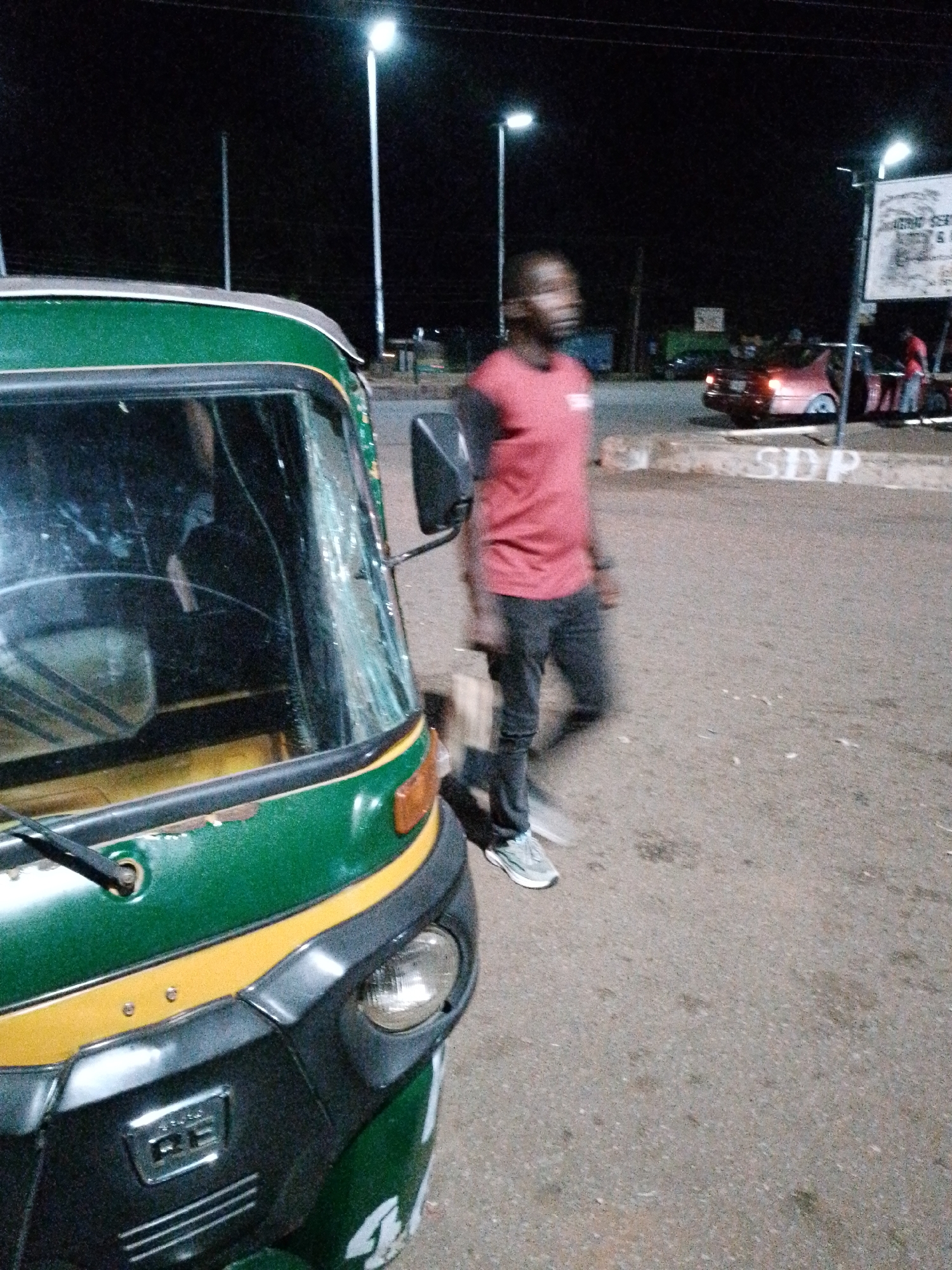 A person is walking in front of a green and yellow auto rickshaw at night. The auto rickshaw has a cracked windshield. The person is wearing a red t-shirt and dark pants. In the background, there is a street light and a red car. A sign with text is visible in the background as well. The ground is paved.