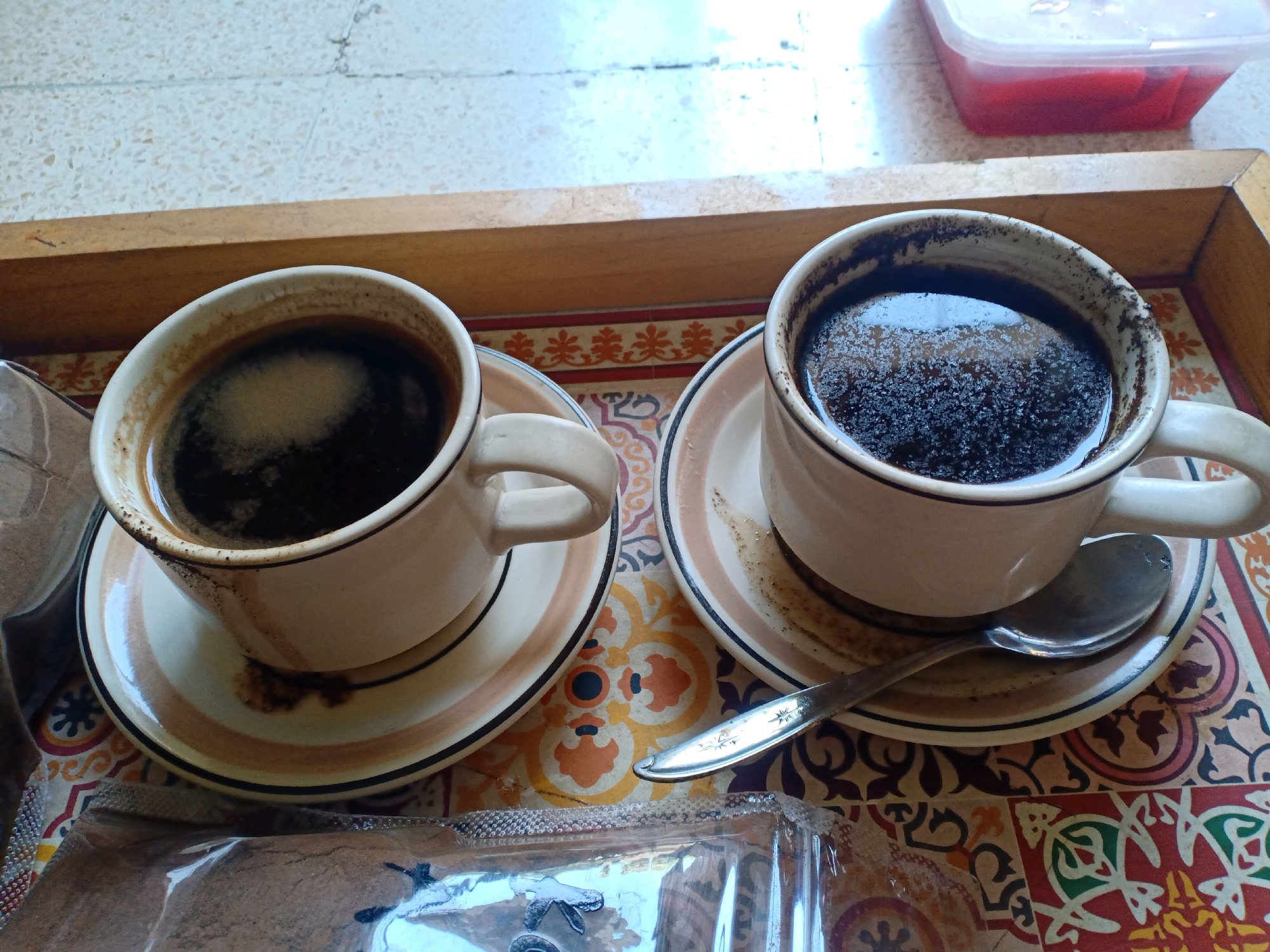 Two cups of dark coffee sit on a tray with a patterned surface. The cups are white with thin colored bands and are filled to the brim with coffee. One cup is to the left, and the other is to the right. A spoon rests on the saucer of the cup on the right. A clear plastic package containing a brown powder, possibly coffee grounds, is visible in the foreground. In the background, a transparent plastic container with red contents is partially visible. The scene appears to be indoors, possibly on a table, with a tiled floor or wall in the background. There is no indication of the specific location within Lumajang, Indonesia.