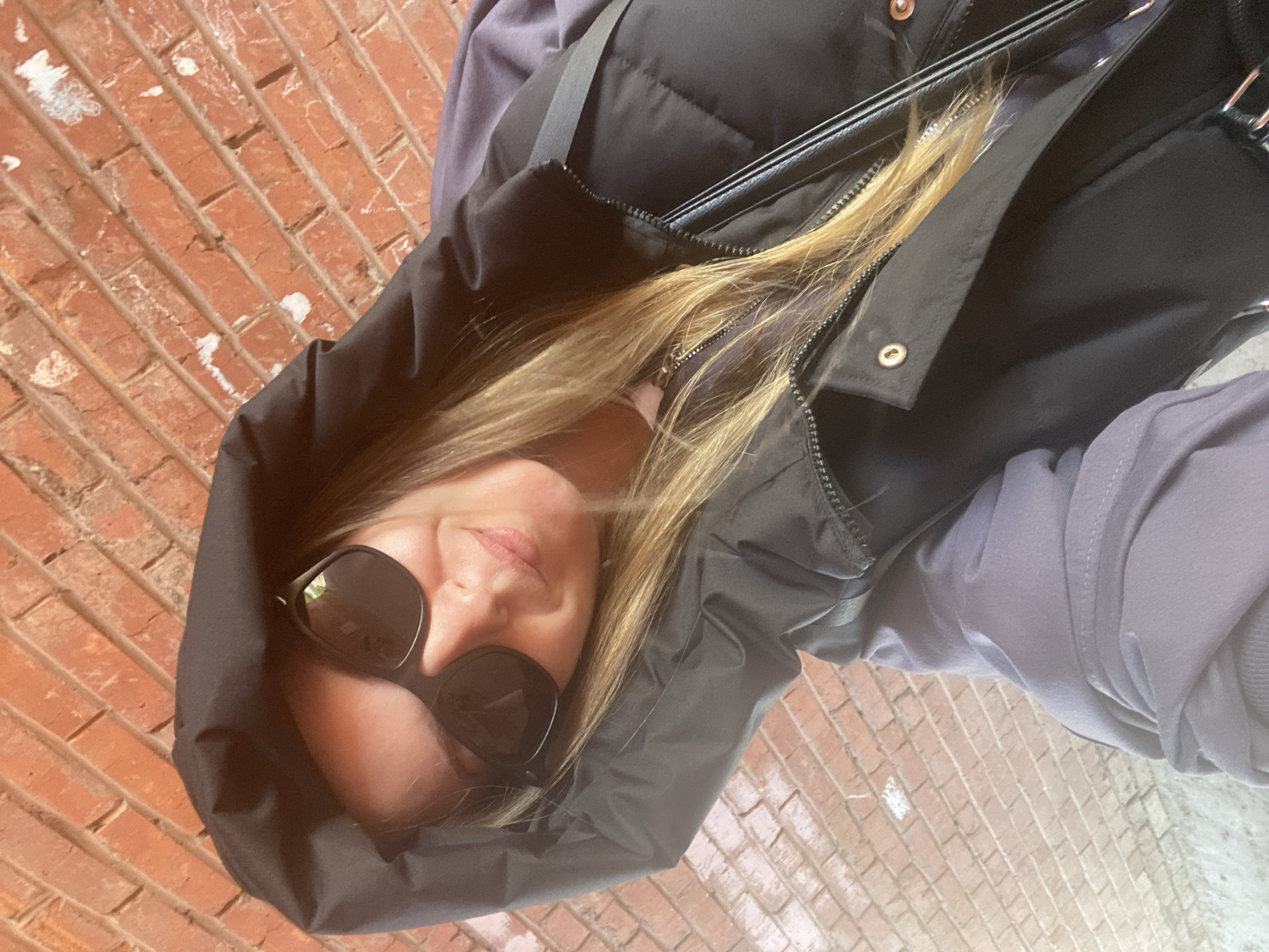 A woman wearing sunglasses and a black hooded vest stands on a red brick sidewalk. Her blonde hair falls around her face and over the vest. A light grey hooded sweatshirt is visible beneath the vest. A black handbag hangs from her shoulder. The bricks of the sidewalk extend into the background. The photo is taken at an angle. It is impossible to determine the location in the city of Noviy, Russia from this image.